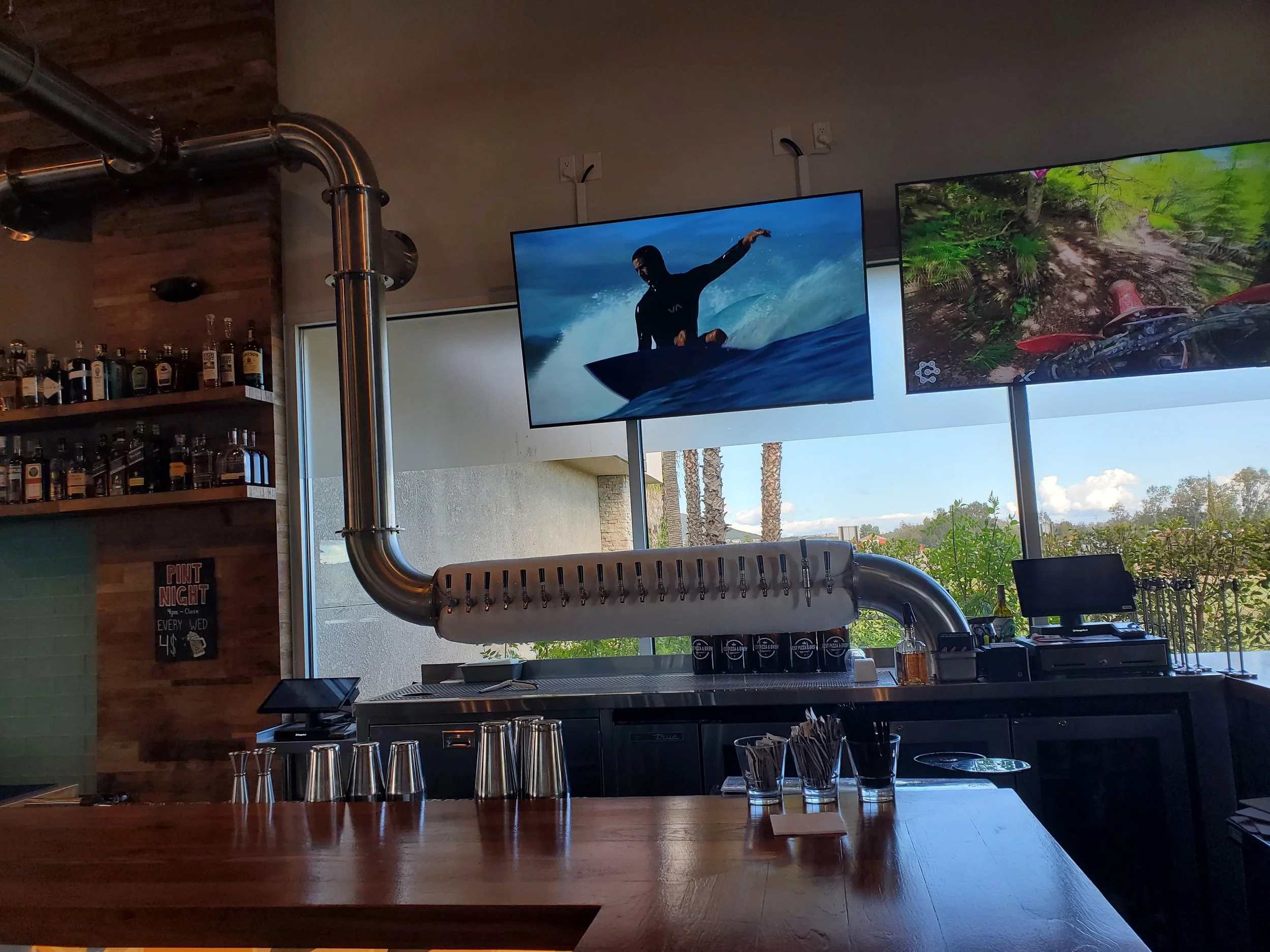 Indoor bar scene with multiple television screens showing surfing and outdoor footage, bar counter with shot glasses on it, and bottles of alcohol on a wooden shelf in the background.