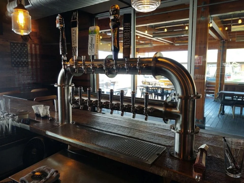 A bar setup with beer taps on a wooden counter in a restaurant or pub, with a window and dining area visible in the background.