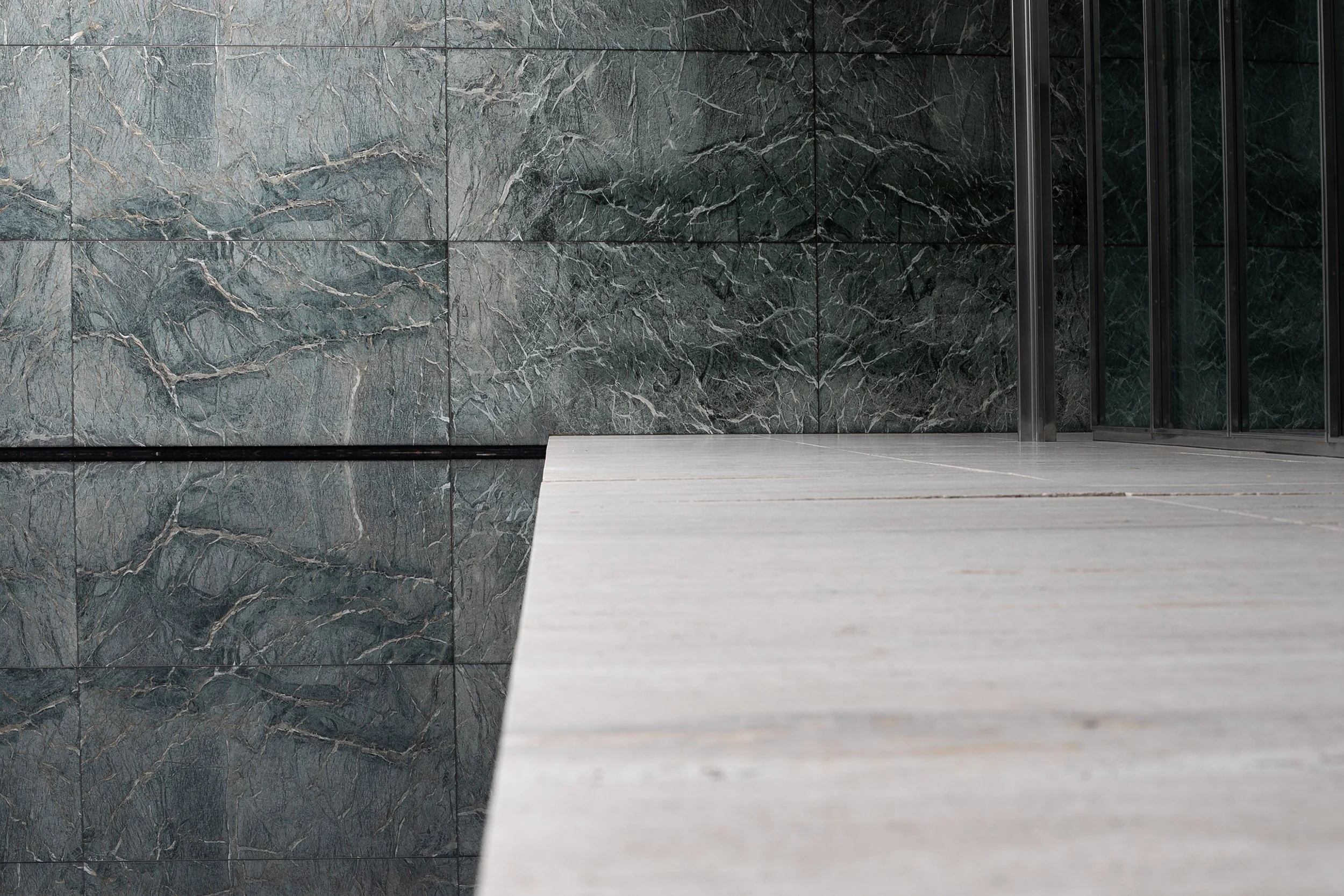 A modern architectural scene featuring a tiled wall with green marble and a smooth concrete walkway.