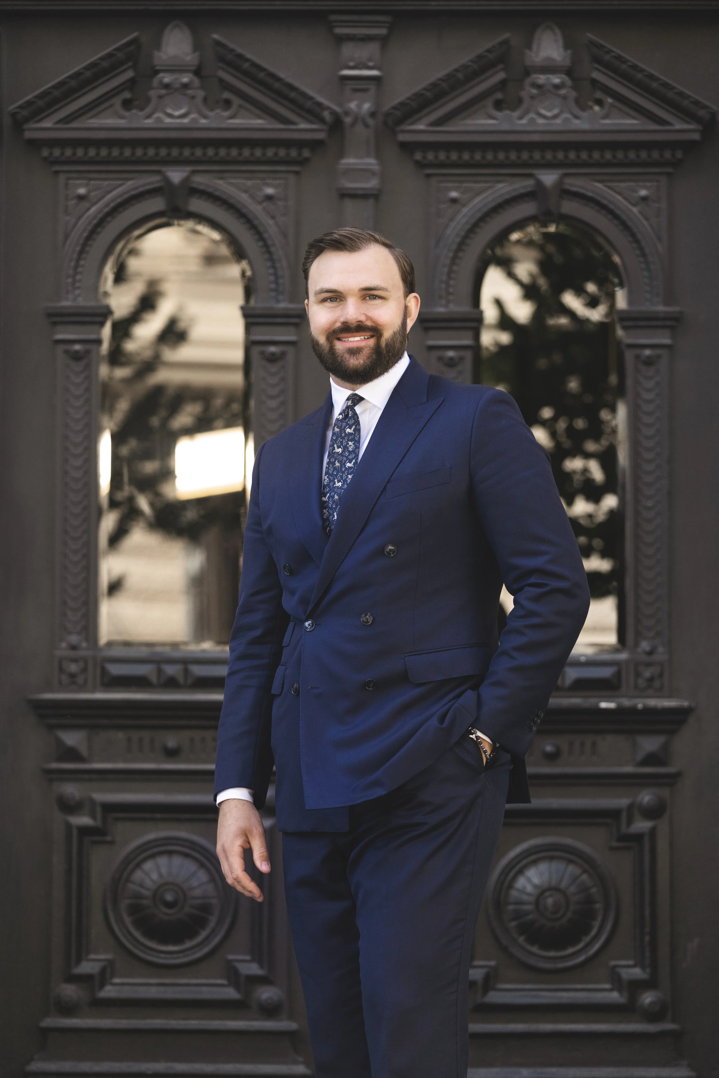 A man in a navy double-breasted suit with a patterned tie standing in front of ornate black doors with arched windows and decorative trim.