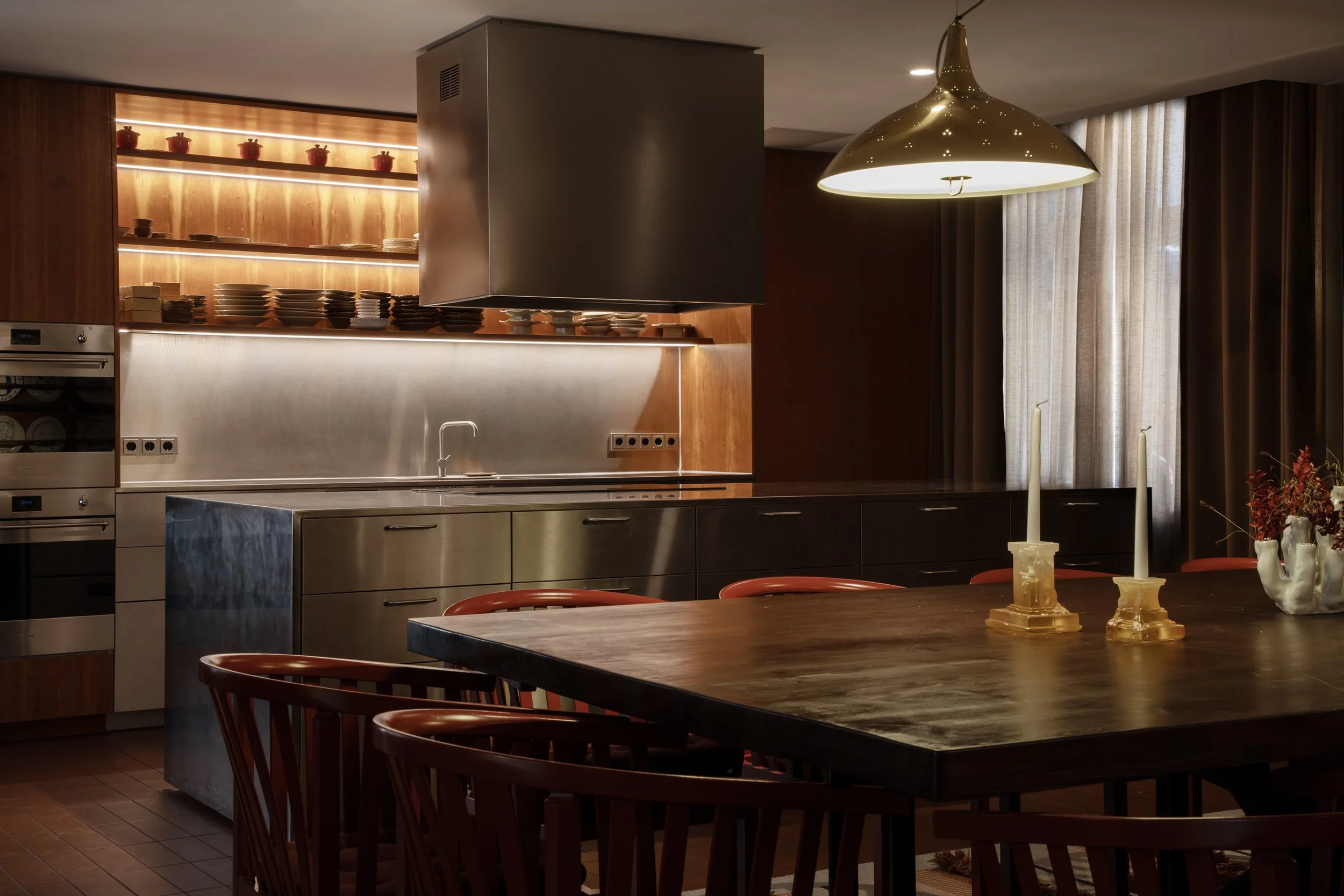 Modern kitchen with wooden paneling, open shelves with dishes, and a large wooden dining table with red chairs. Candles and a vase with flowers decorate the table, illuminated by a pendant light.