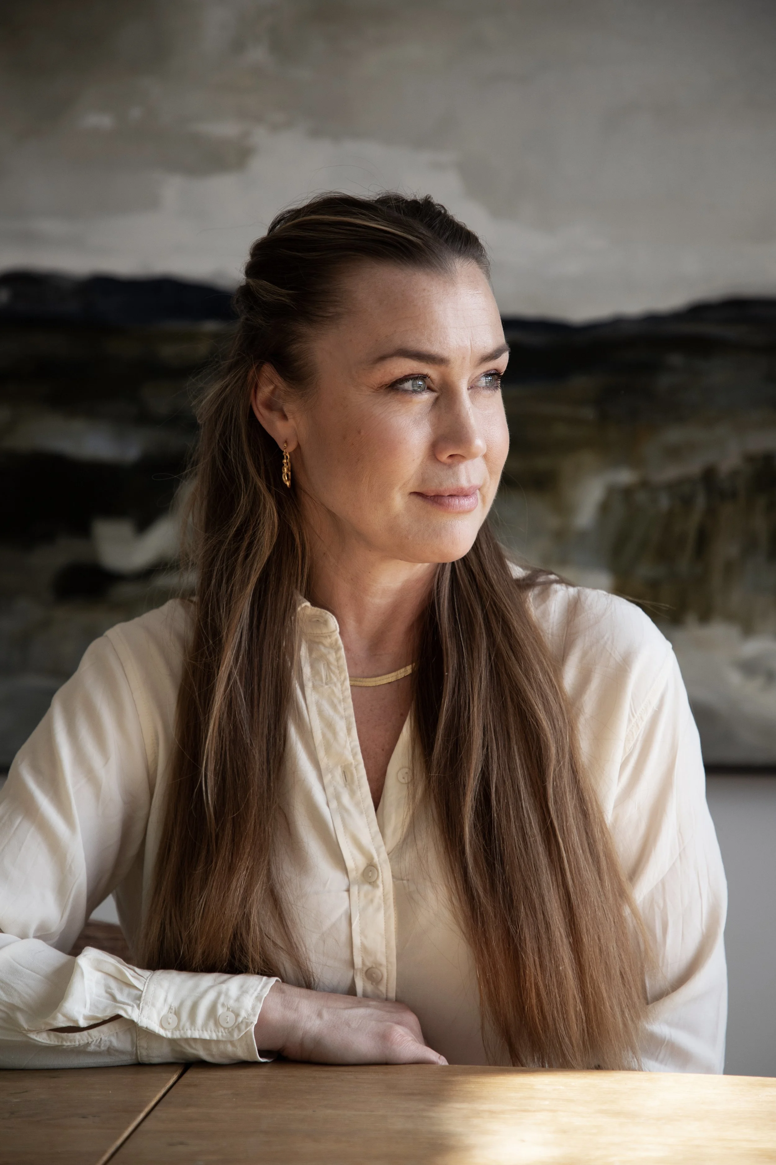 A woman with long brown hair wearing a cream-colored blouse and gold jewelry, sitting at a wooden table and looking to the side with a thoughtful expression.