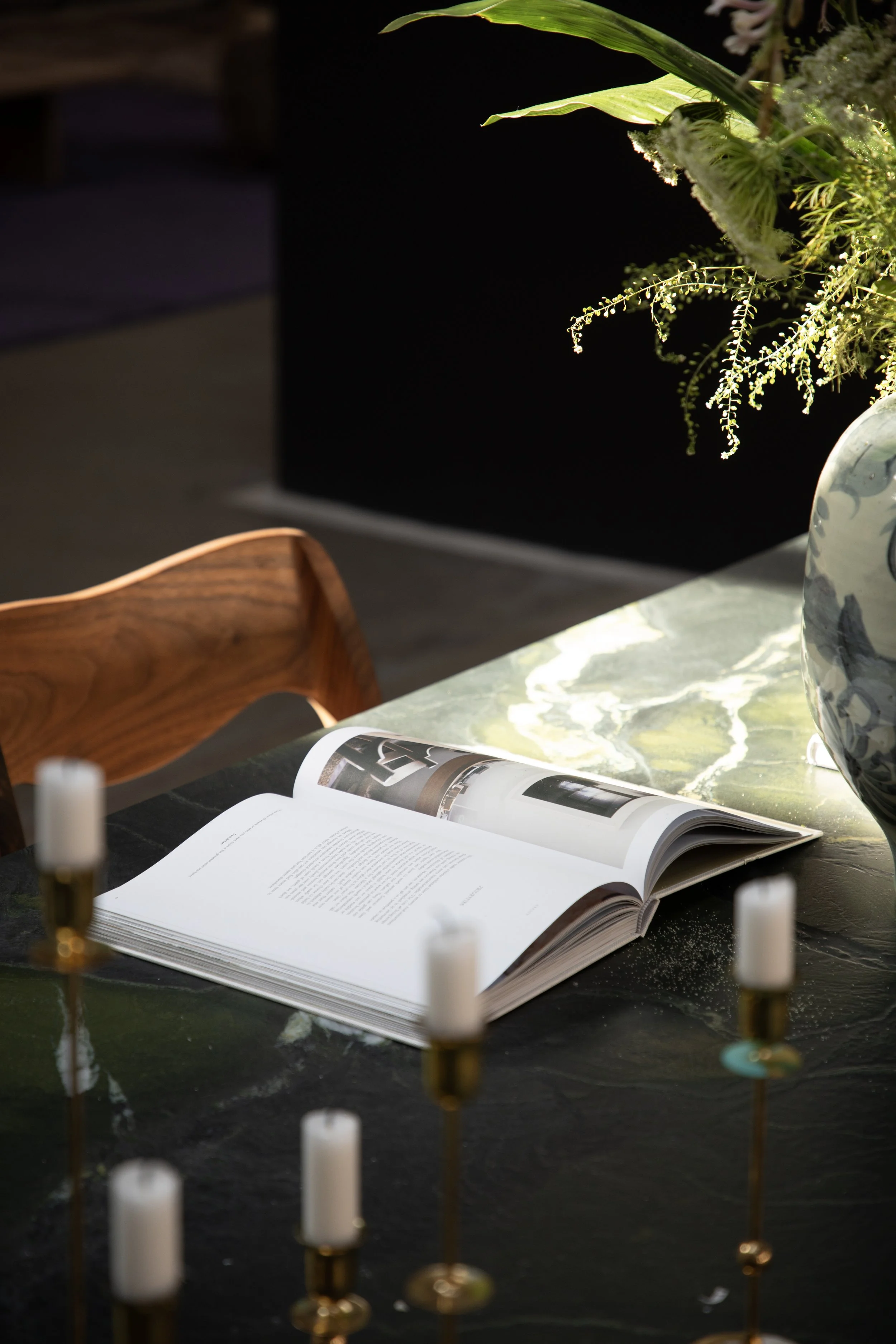 An open magazine on a black marble table, with white candles in brass holders and a large flower vase with green foliage nearby.