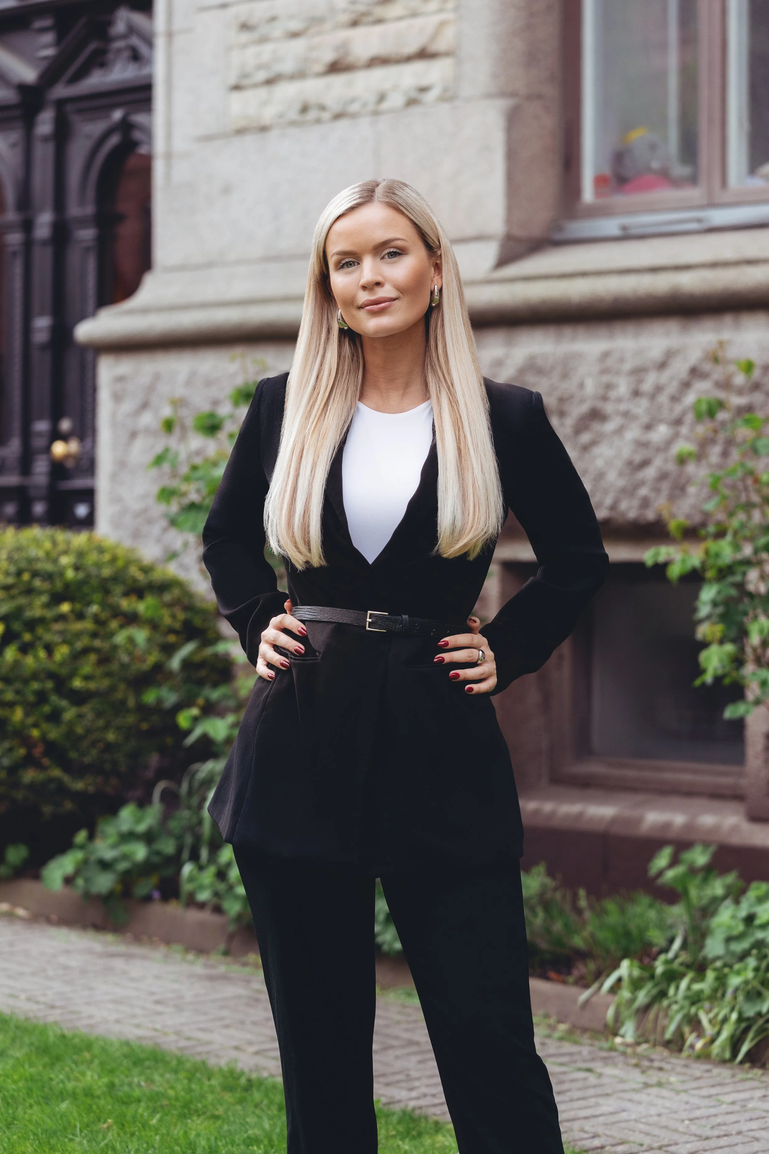 A woman with long blonde hair wearing a black suit with a white top, standing outside in front of a stone building with greenery.