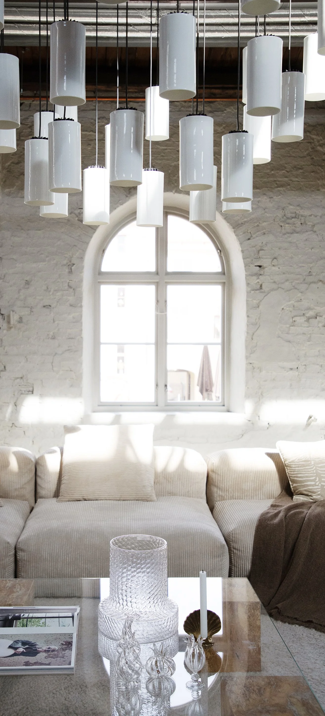 Modern interior with white brick wall, large arched window, beige sofa, glass coffee table with decorative glass vases, and cylindrical pendant light fixtures hanging from ceiling.