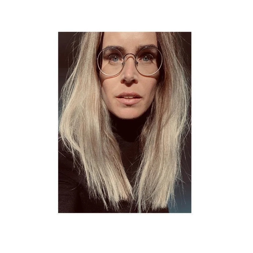 A woman with long blonde hair wearing round glasses and a black top, looking directly at the camera indoors.