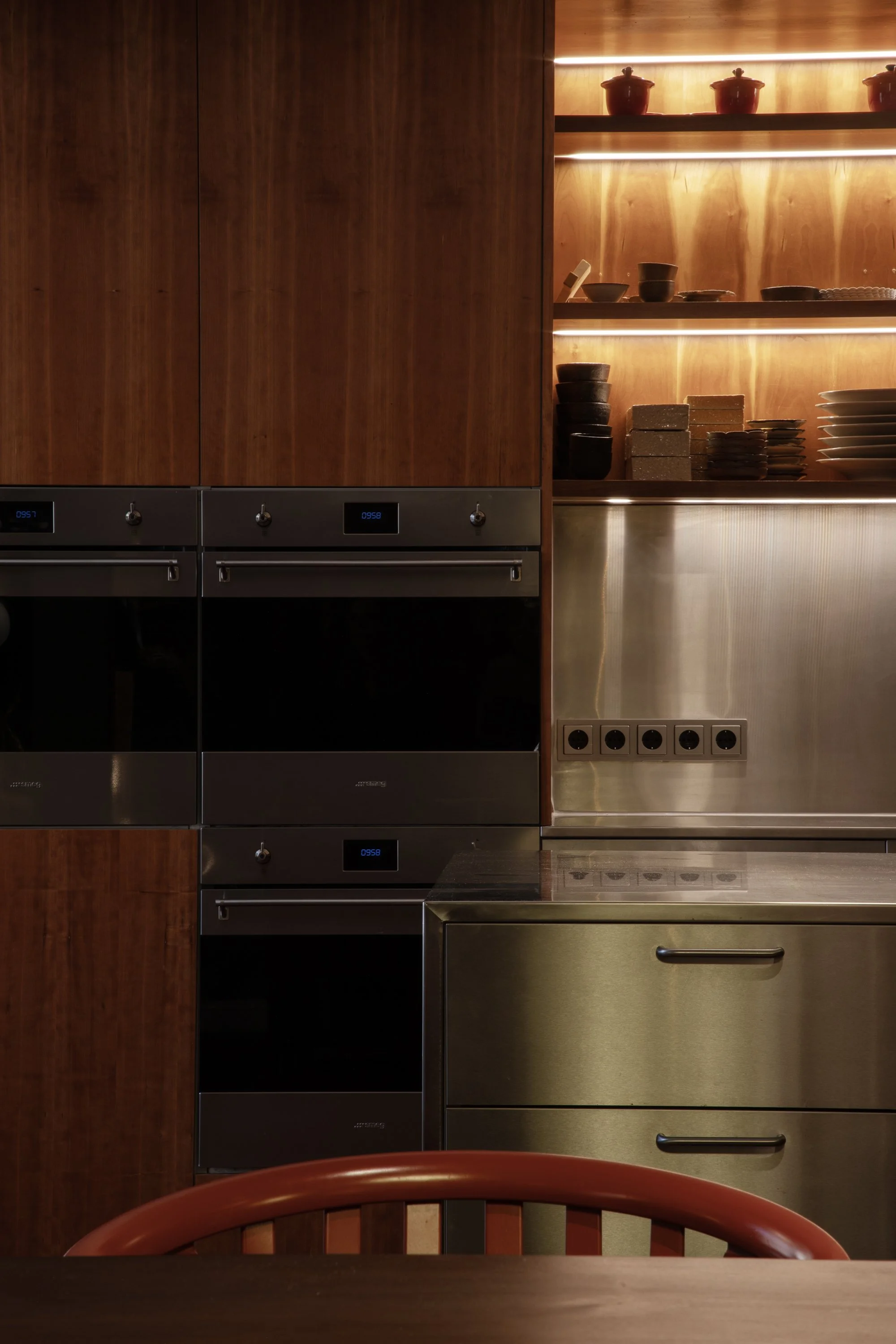 Modern kitchen with wooden cabinets, black oven and microwave, and open shelves with bowls and plates; a wooden chair with a curved backrest is partially visible at the bottom.