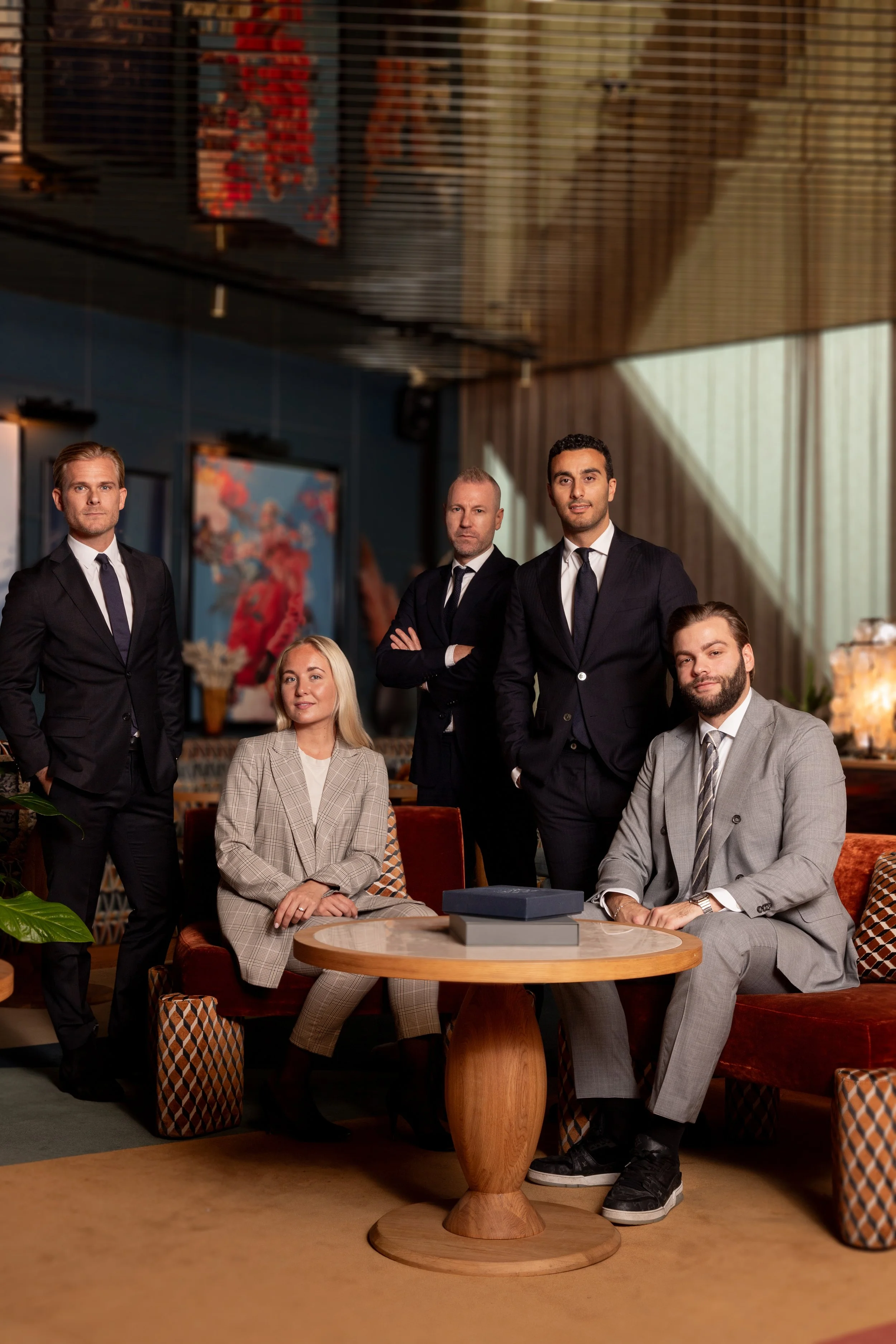 A group of five professionally dressed people, four men and one woman, in a modern, stylish room with warm lighting, some seated and others standing, with abstract artwork on the wall behind them.