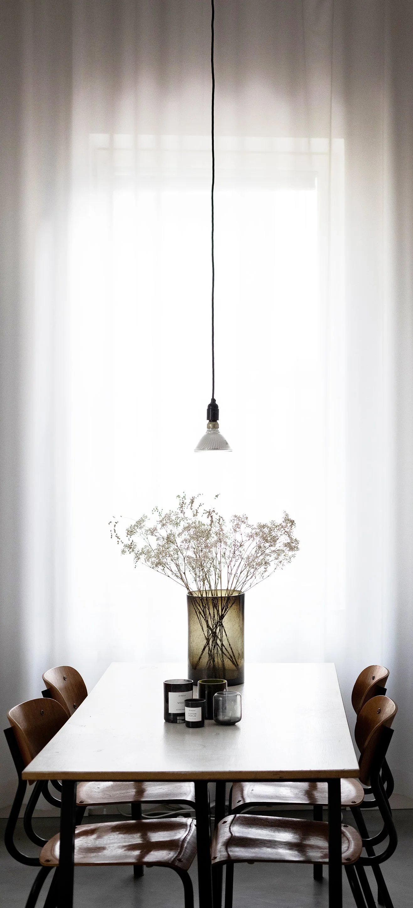 Dining room with a white table, surrounded by six wooden chairs, a large vase with dried flowers, and five candles on the table. A hanging light with a black cord is above the table, and sheer curtains cover a bright window in the background.