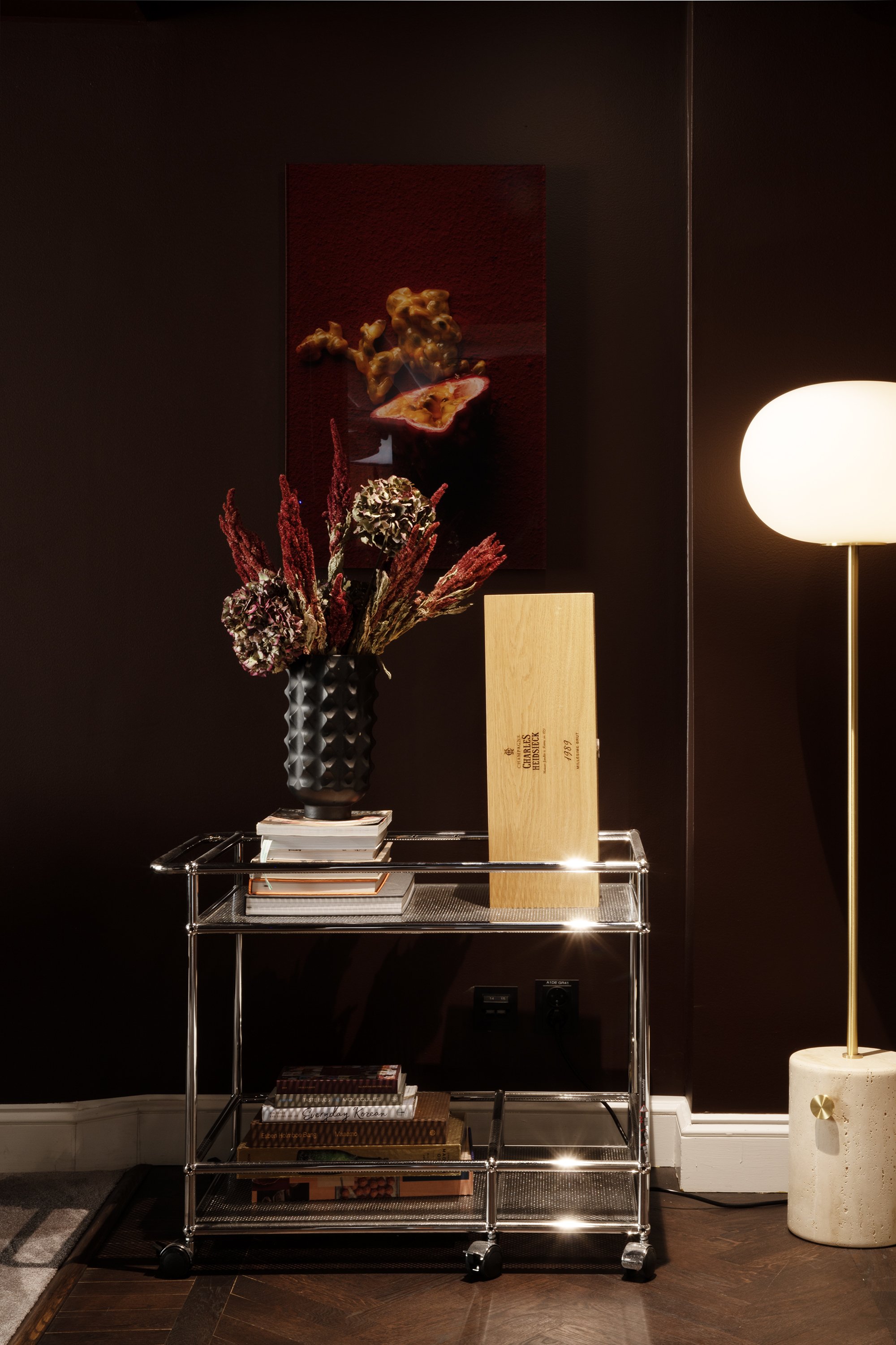 A modern interior with a dark wall, illuminated by a floor lamp. A glass cart holds a black textured vase with dried, red and green flowers, and a wooden box labeled 'Charles Heidsieck.' There are books and magazines on the lower shelves. A framed art piece with a 3D effect hangs on the wall.