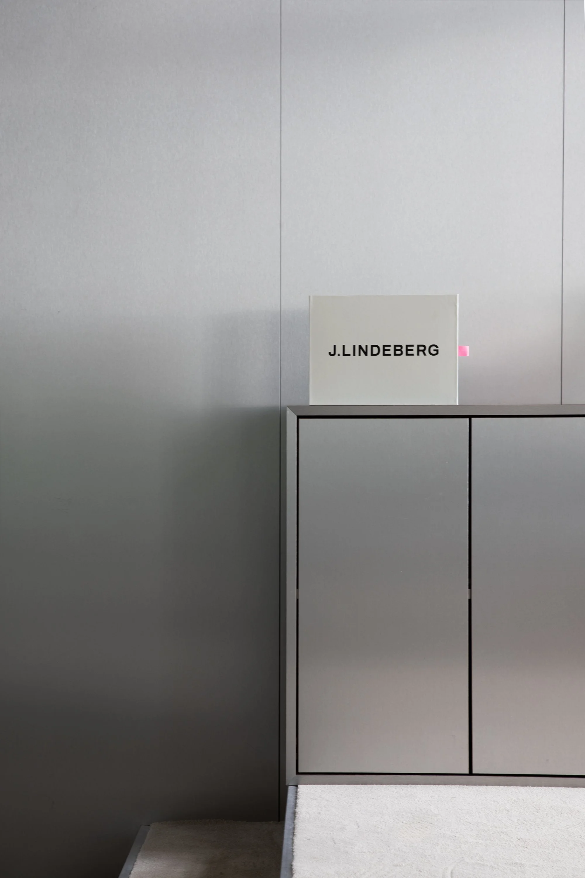 A minimalistic modern reception area with a gray cabinet and a white sign that says 'J. LINDEBERG' in black letters, set against a plain gray wall.
