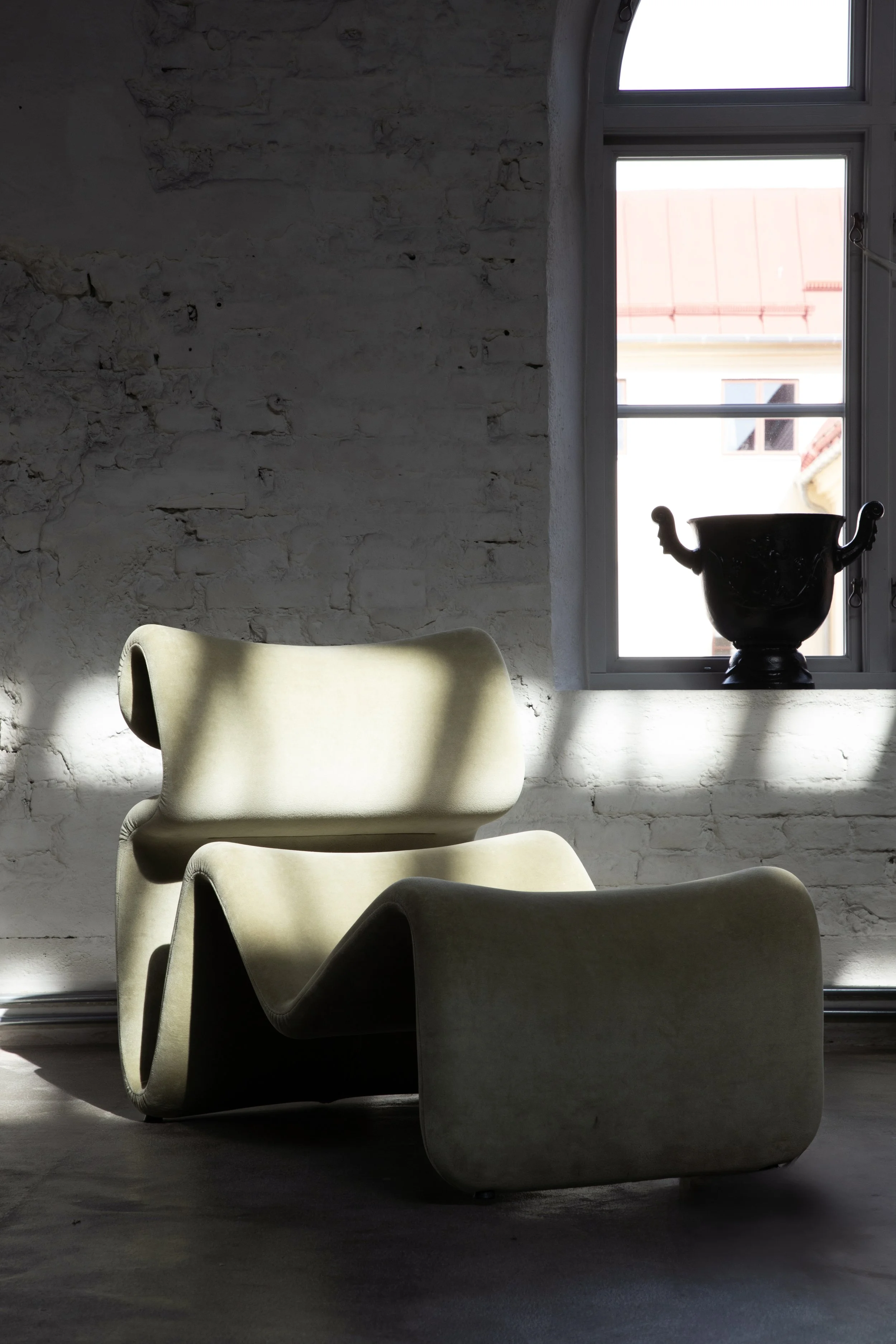 Modern beige lounge chair in a room with white brick walls, a window with sunlight streaming in, and a black vase on the window sill.