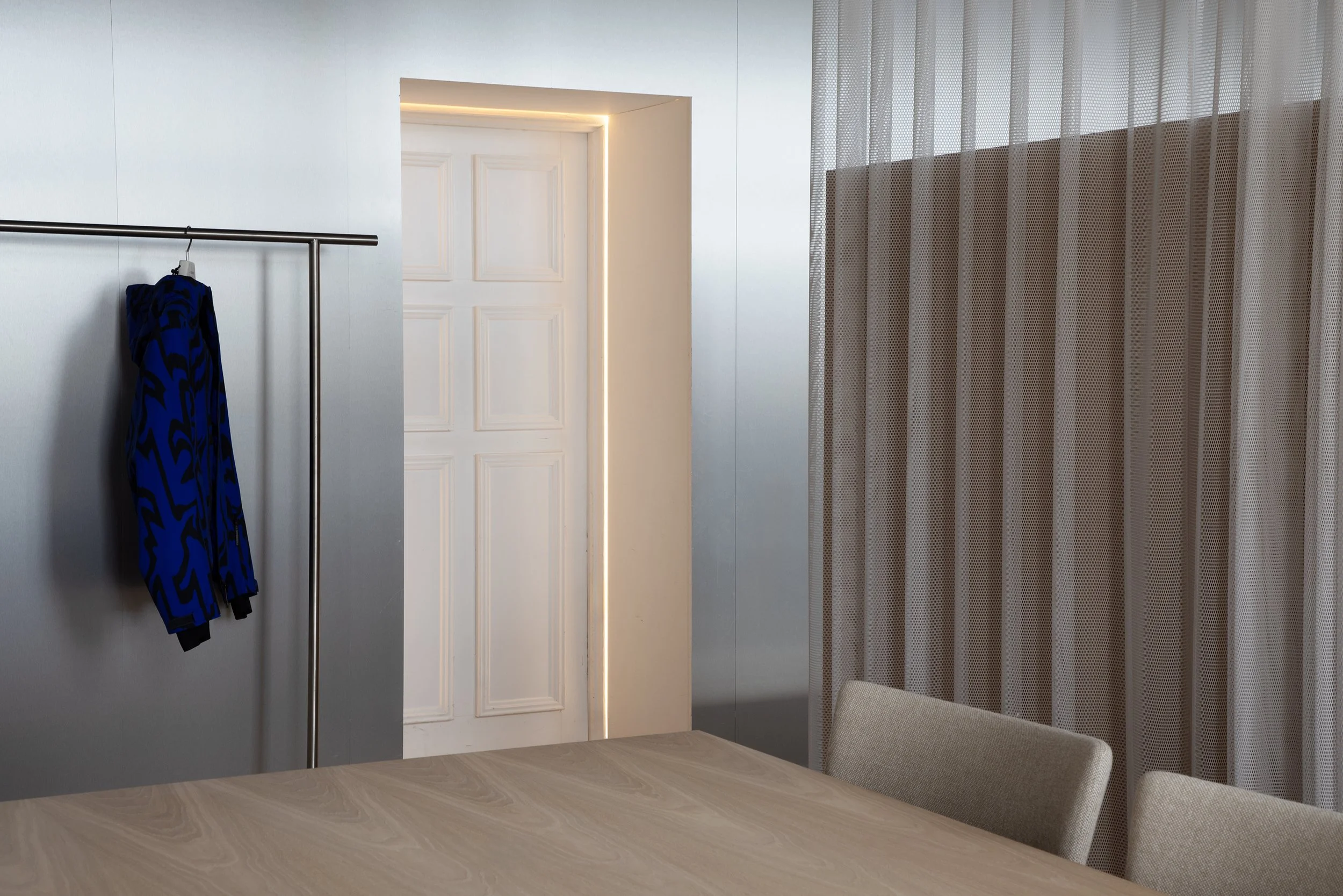 Modern interior with a door, a clothing rack with a blue and black patterned garment, beige chairs, curtains, and a light-colored wooden table.