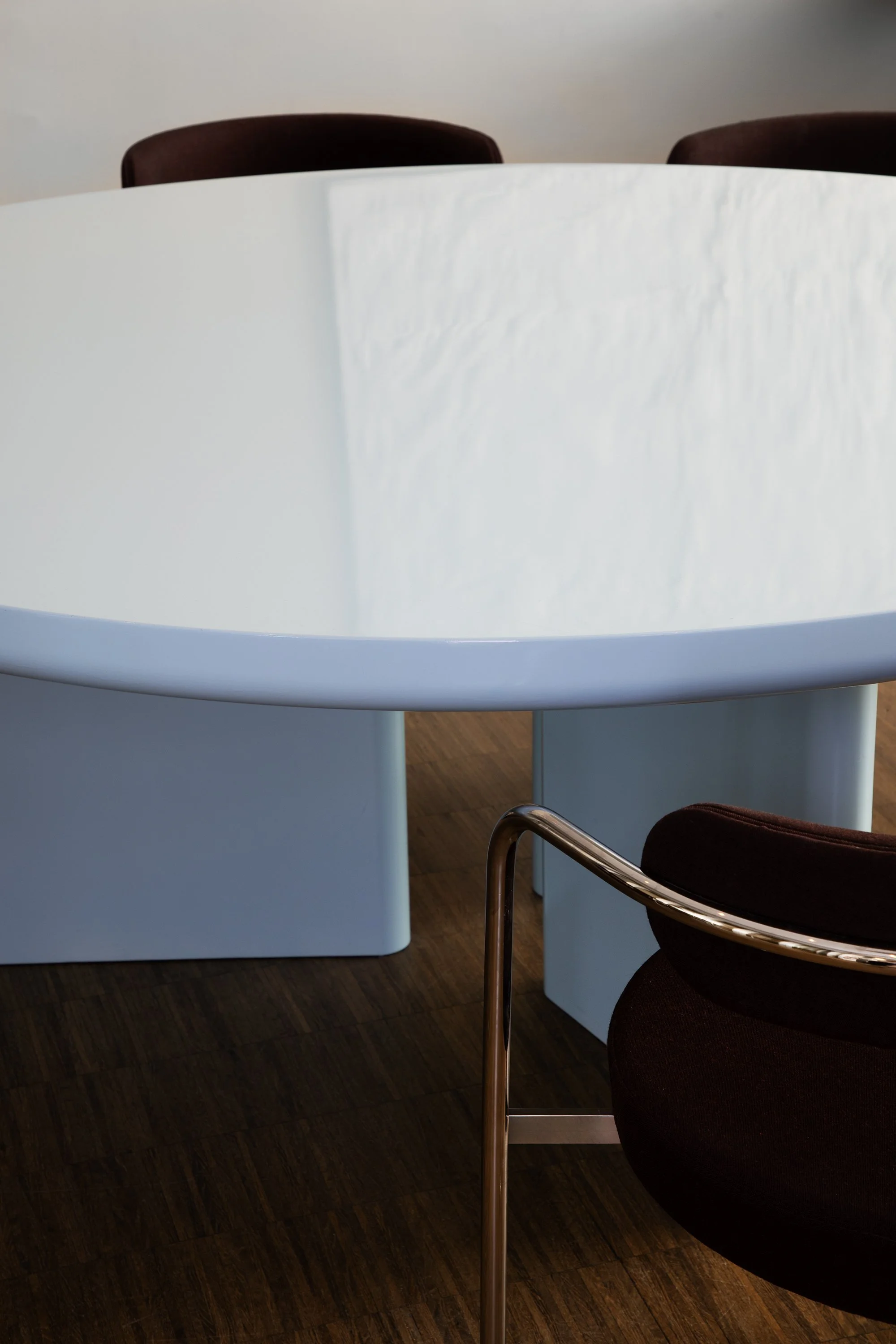 Close-up of a modern round table with a white top and a blue base, surrounded by dark brown chairs with metal arms, in a room with wooden flooring.