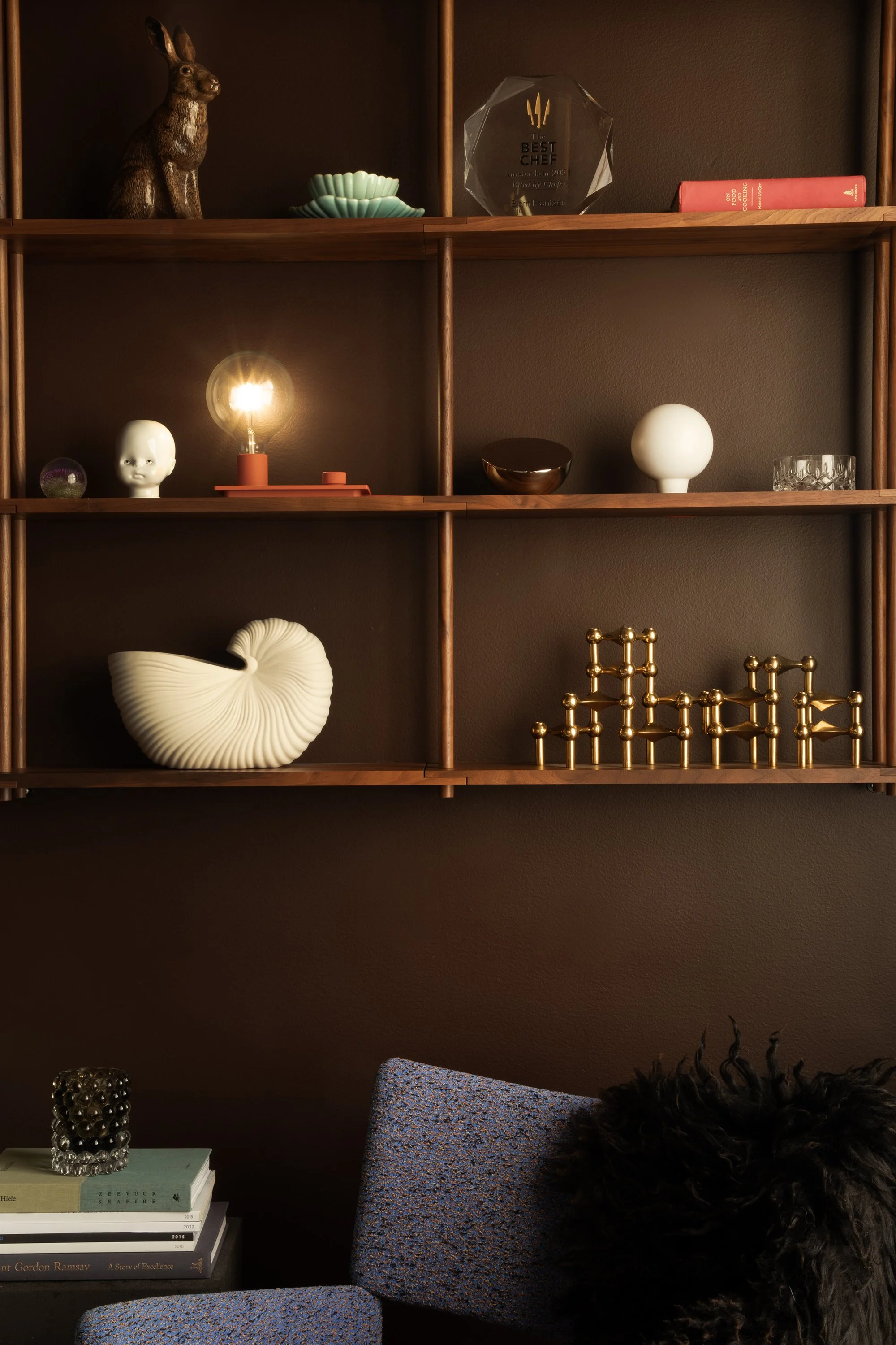 A wooden wall shelf with various decorative objects, including a ceramic rabbit, a small sculpture, a lamp, a ceramic head, a black bowl, a white sphere, a glass, and gold geometric sculptures. Part of an upholstered chair with a black furry cushion and some books are visible at the bottom.