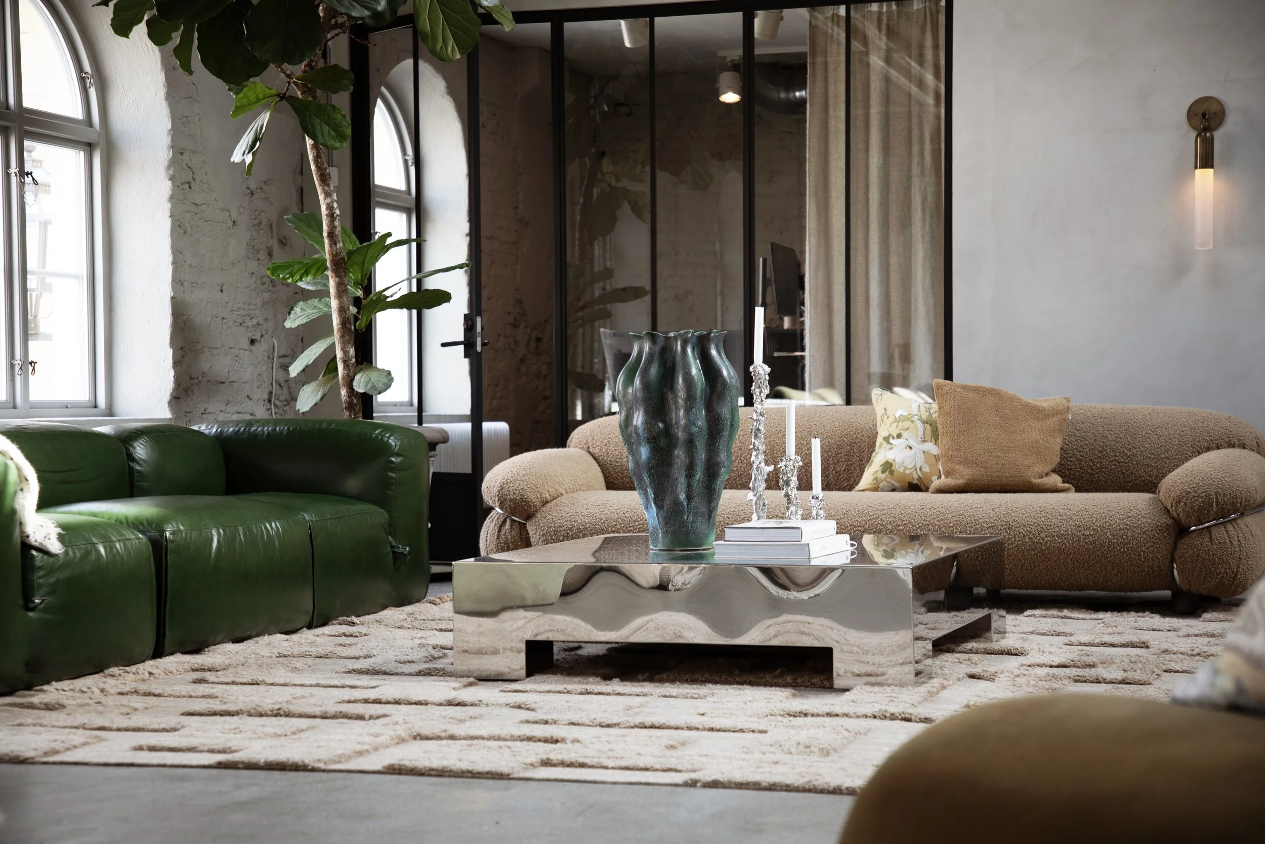 Modern living room with beige and green sofas, a mirrored coffee table, decorative vases and candles, large windows with natural light, and a tall indoor plant.