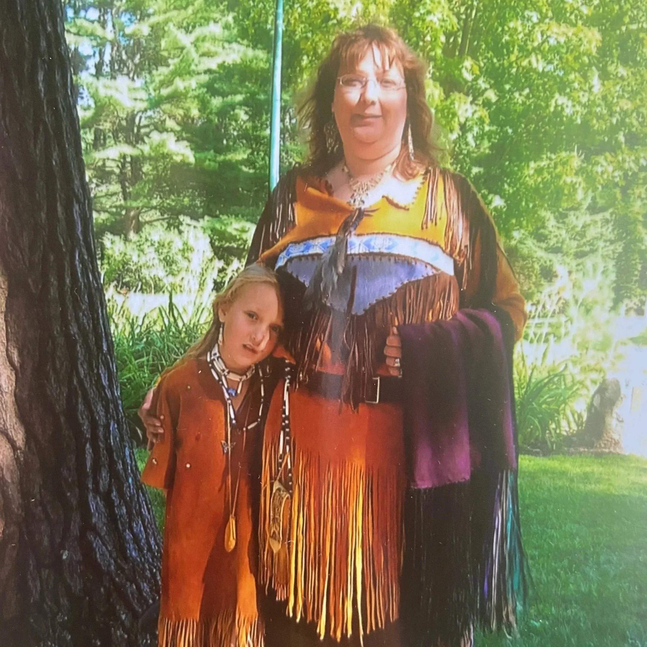 A woman and a young girl dressed in Native American-inspired clothing standing outdoors with greenery and trees in the background.