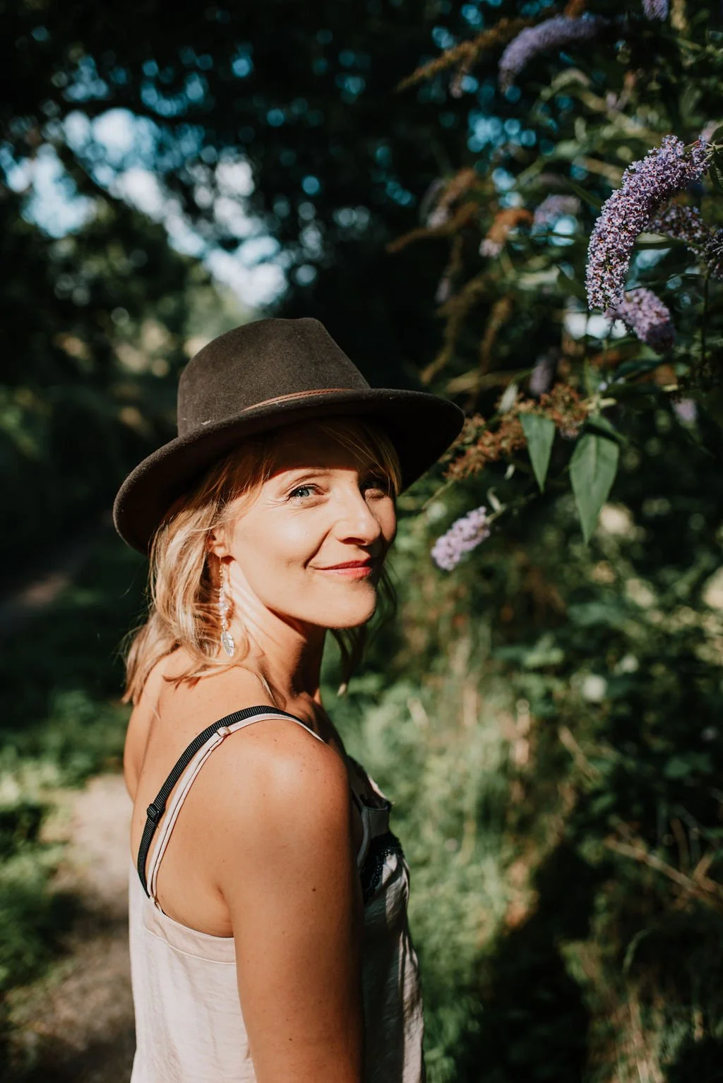 A woman wearing a brown hat and earrings standing outdoors among greenery and purple flowers, smiling slightly at the camera.