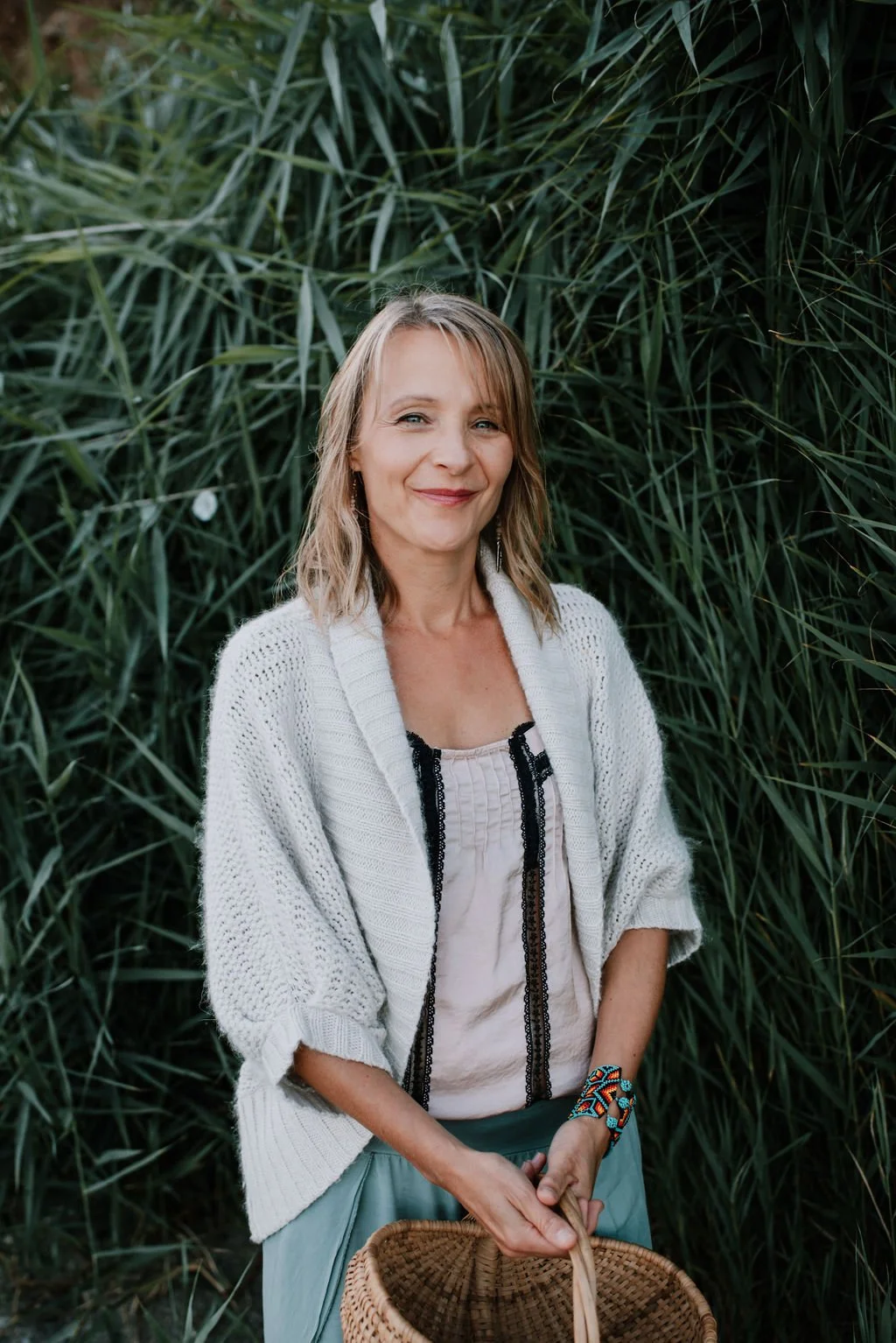 Catherine Hale — woman in a cream knit cardigan holding a wicker basket, smiling in front of tall green reeds — nervous system integration and embodied leadership.