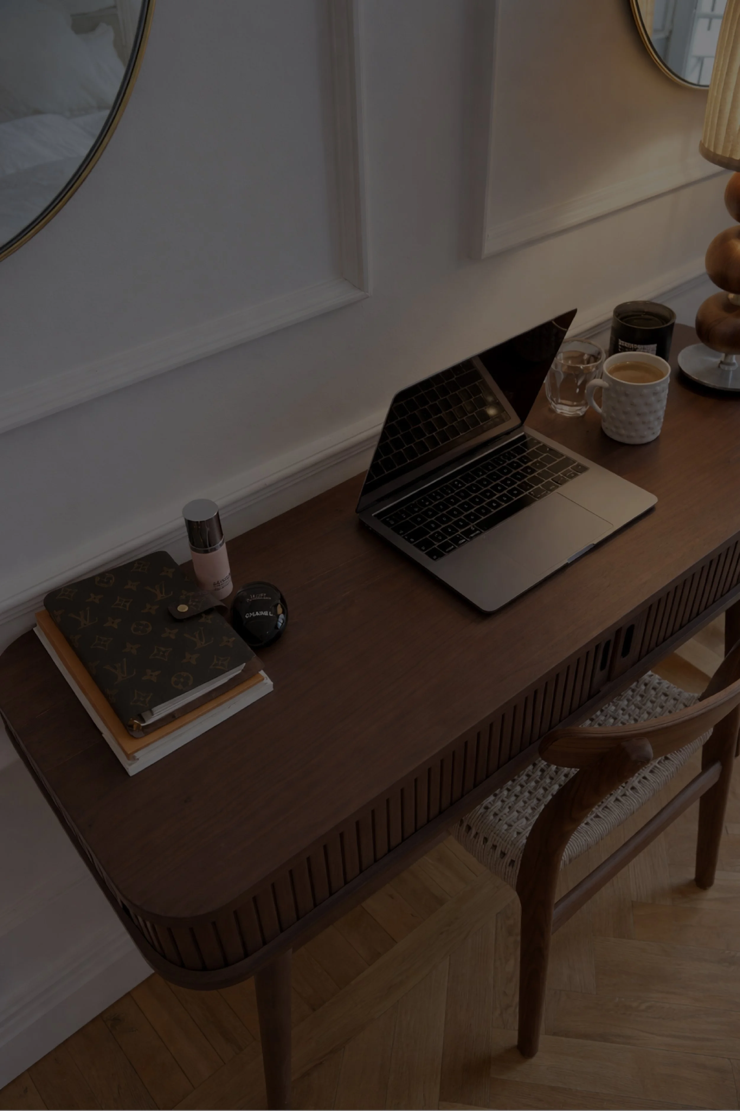 A wooden desk with a silver laptop, a mountain of two notebooks, lip balm, a coaster with a container on it, a glass of water, a mug with coffee, a small candle, and a decorative lamp with a beige shade. The desk is against a wall with two round mirrors and a chair with a woven seat.