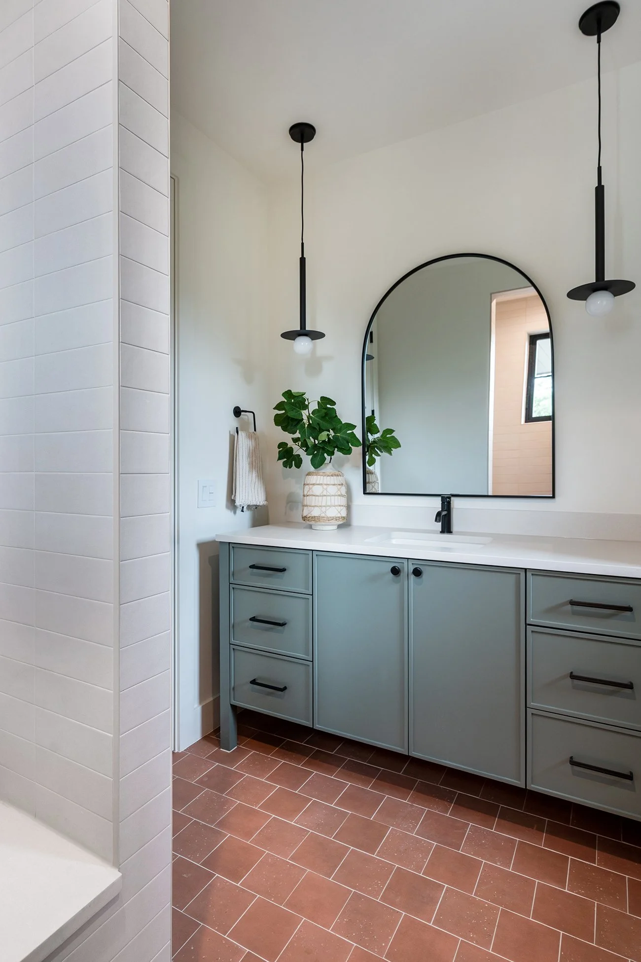 Modern guest bathroom in an Austin Northwest Hills home featuring a muted green vanity, terracotta tile flooring, arched mirror, black fixtures, and clean white walls.