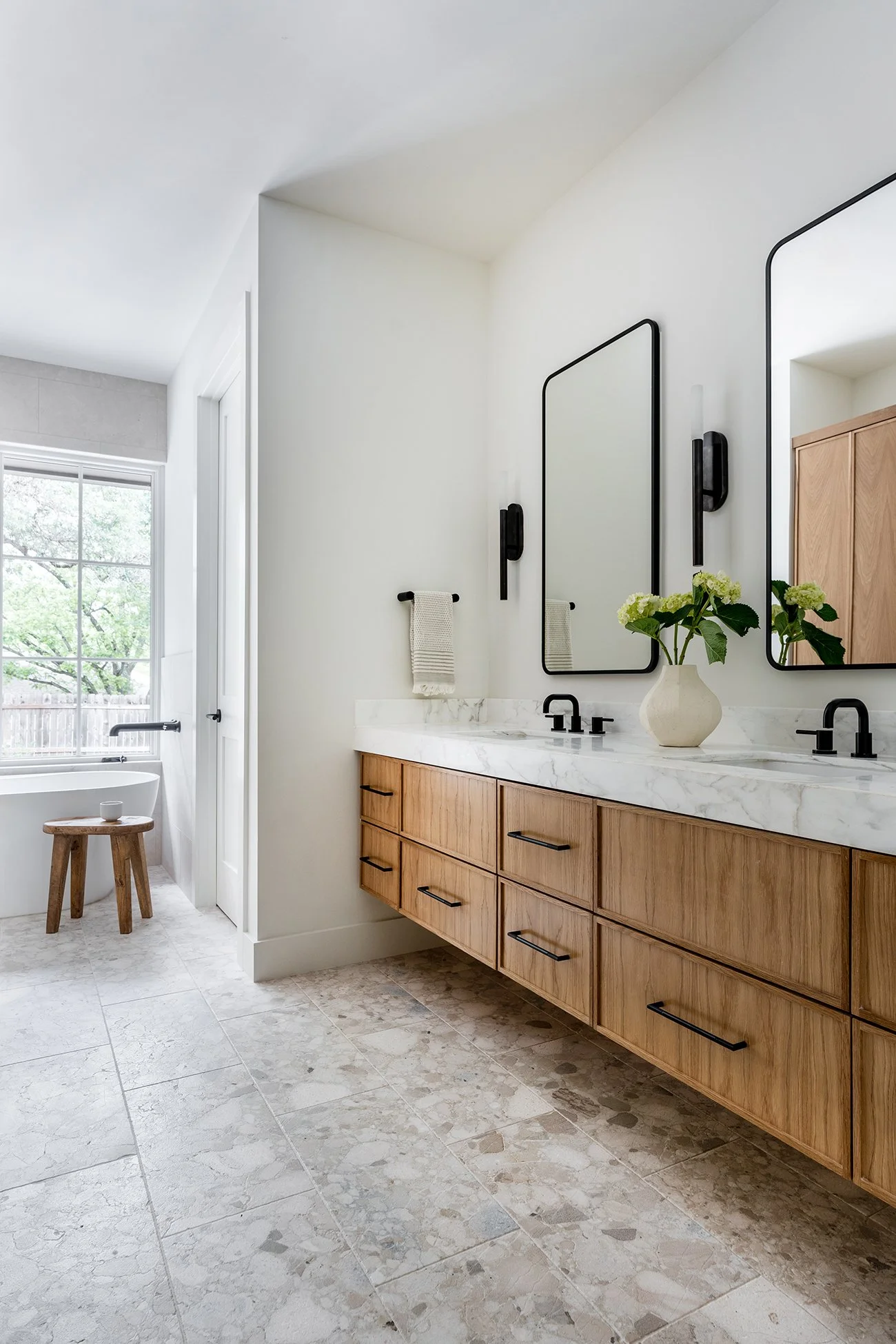Light-filled primary bathroom in a Northwest Hills Austin new build featuring a custom wood double vanity, marble countertop, black fixtures, oversized mirrors, and natural stone flooring.