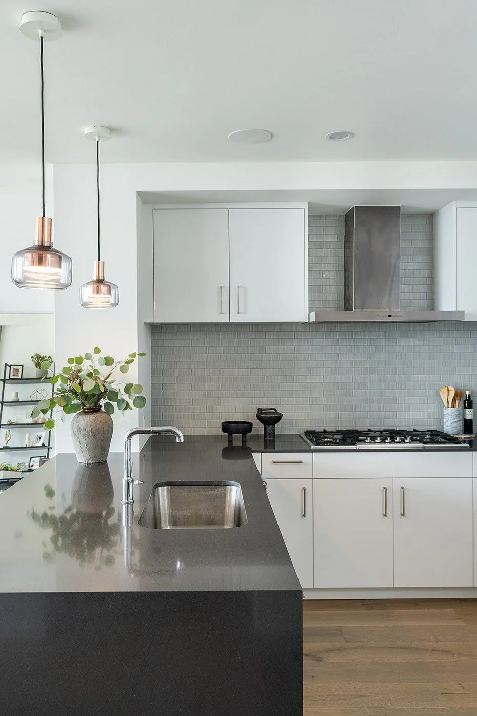 Modern kitchen with white cabinets, stainless steel range hood, gray tile backsplash, black countertop, and pendant lights with glass shades.