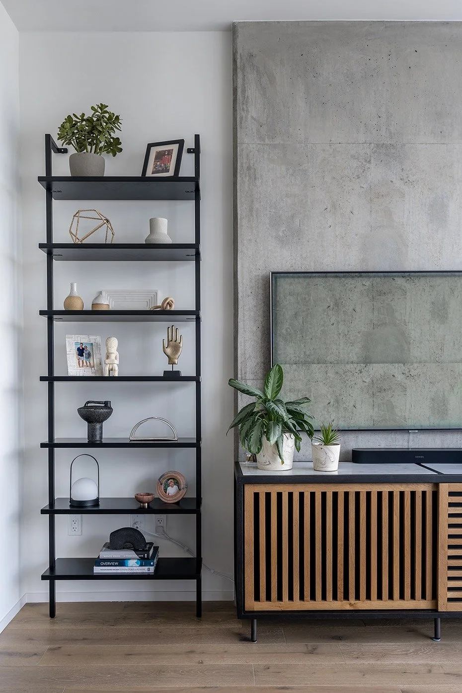 A living room corner featuring a black metal open-shelf with decorative items, and a TV stand with two potted plants, a large wall-mounted TV, and a concrete accent wall.