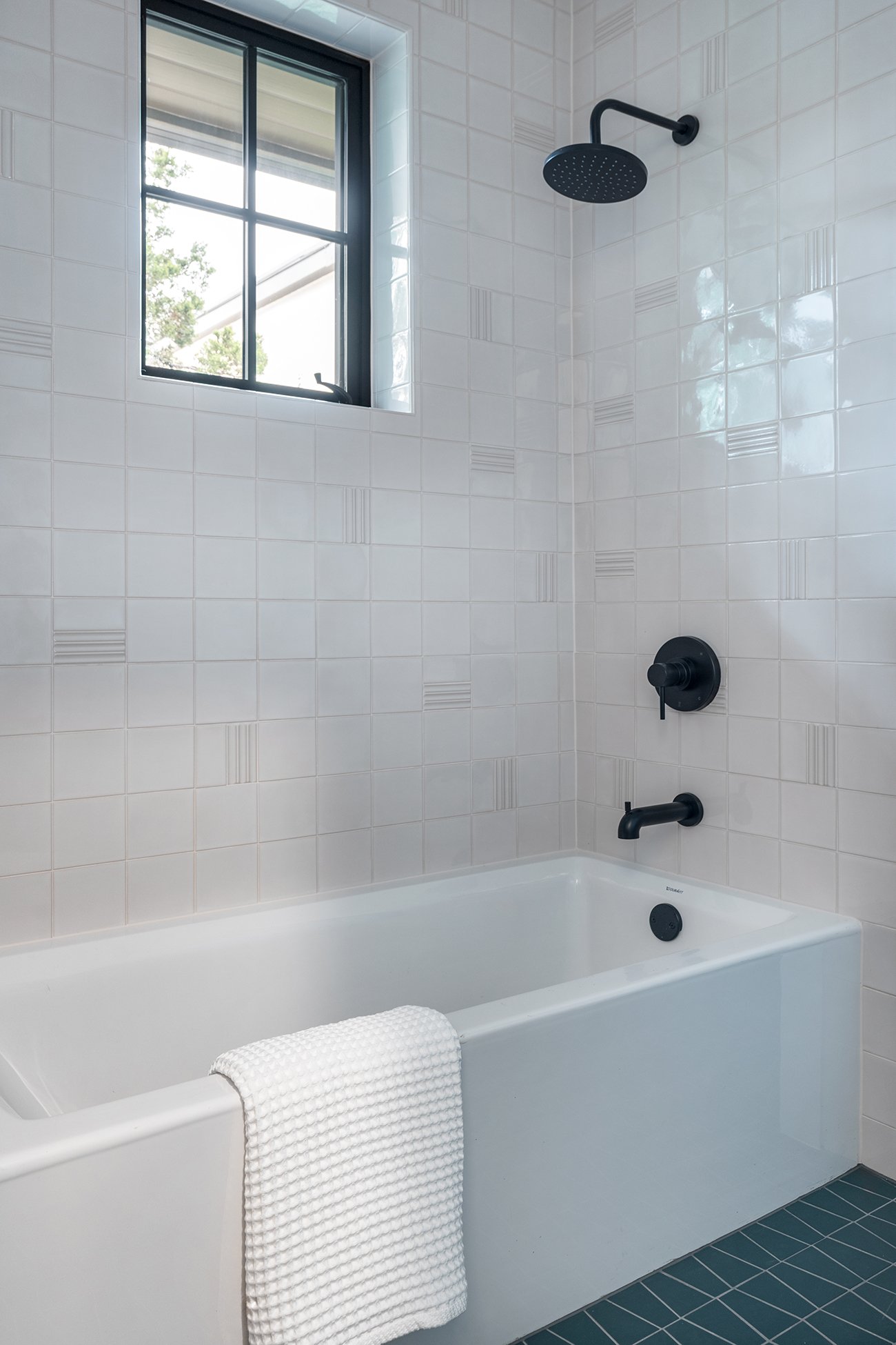 Kids’ bathroom tub and shower in an Austin Northwest Hills home featuring white textured tile, teal floor tile, black plumbing fixtures, and a black-framed window.
