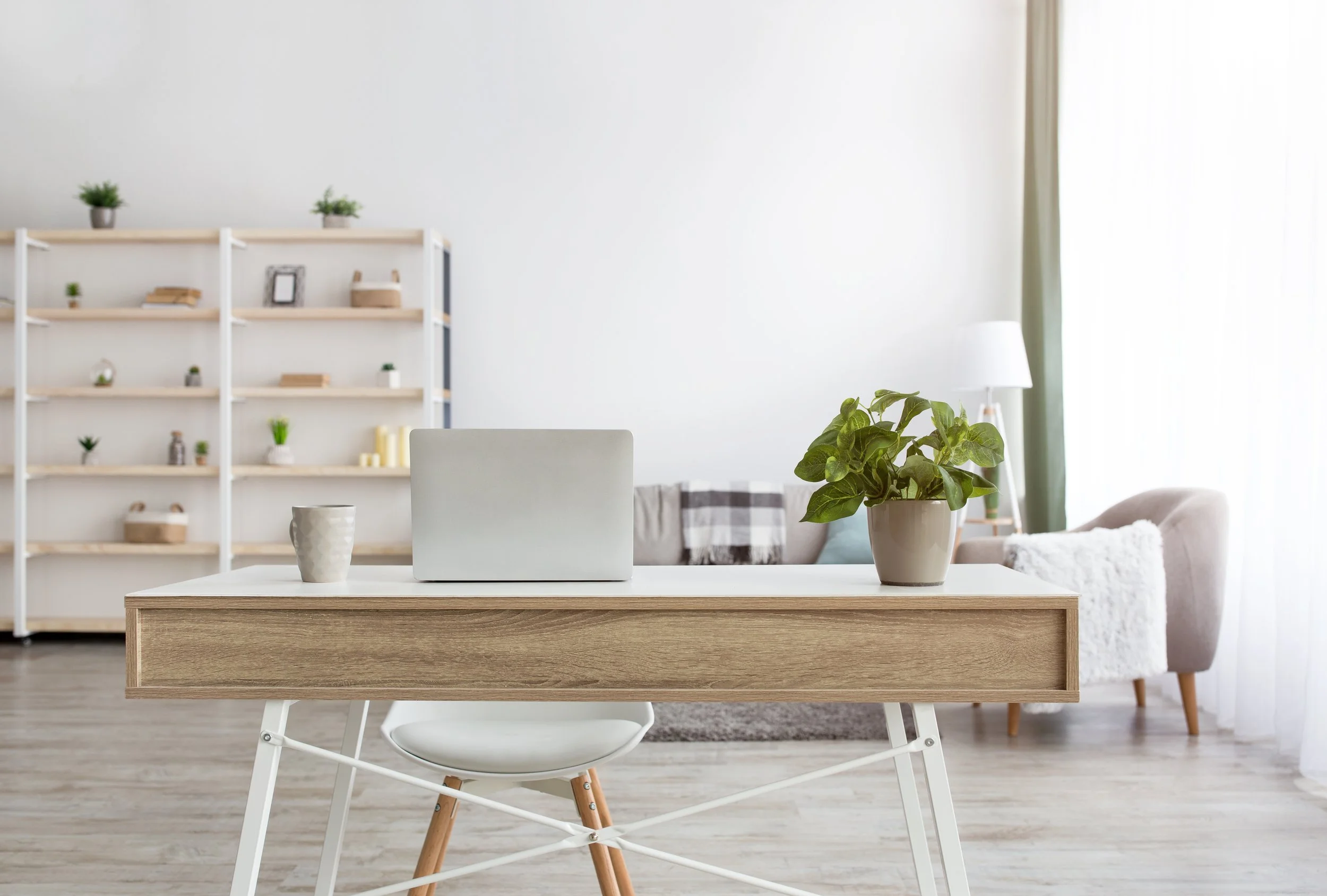 Bright and airy home office with a light wooden desk, a laptop, a potted plant, a mug, and a modern white chair. In the background, there is a cozy sofa with cushions, a shelf with decorative items, and natural light coming through sheer curtains.
