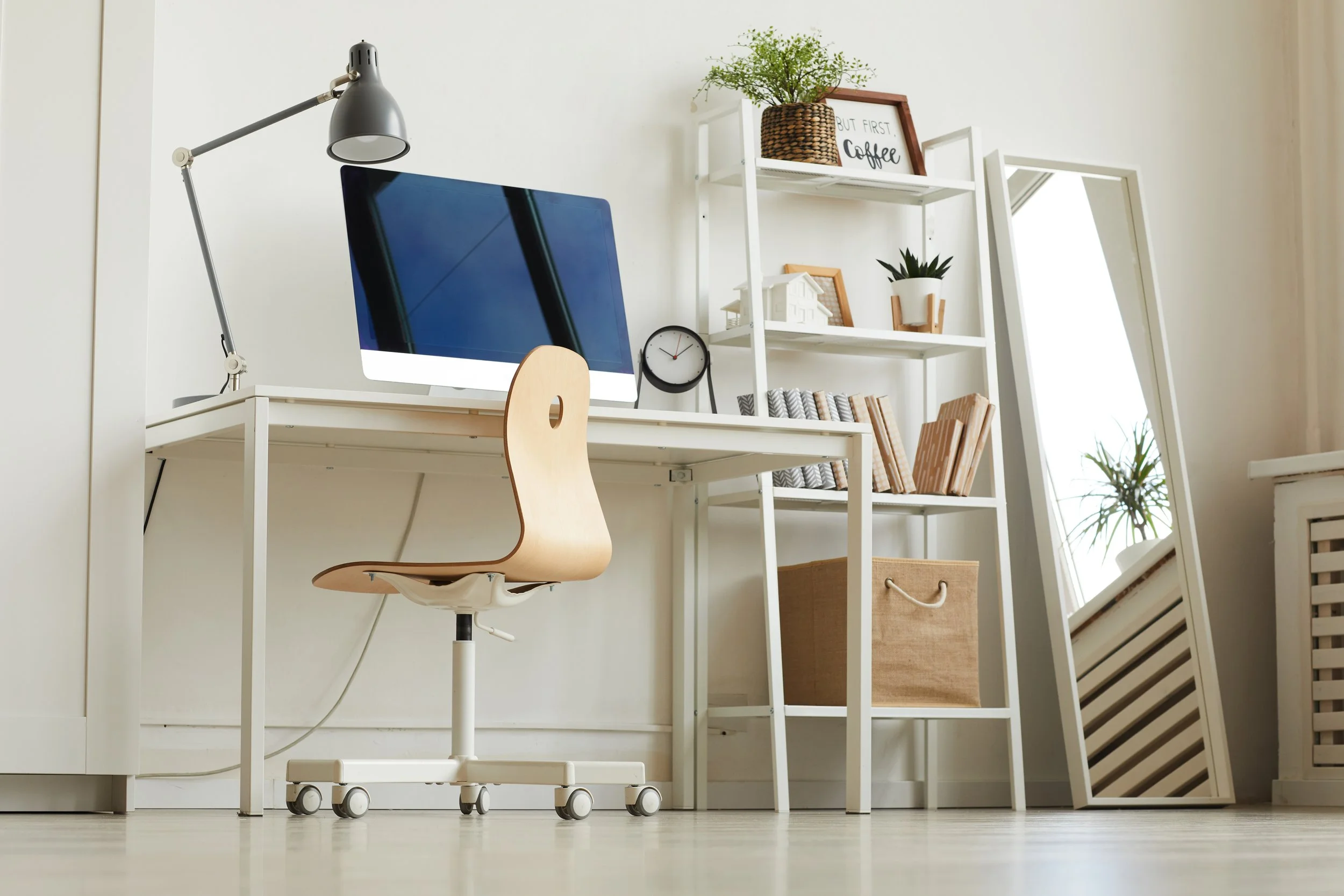 Home office setup with white desk, computer monitor, wooden swivel chair, white bookshelf with decorative items, potted plants, and a full-length mirror leaning against the wall.