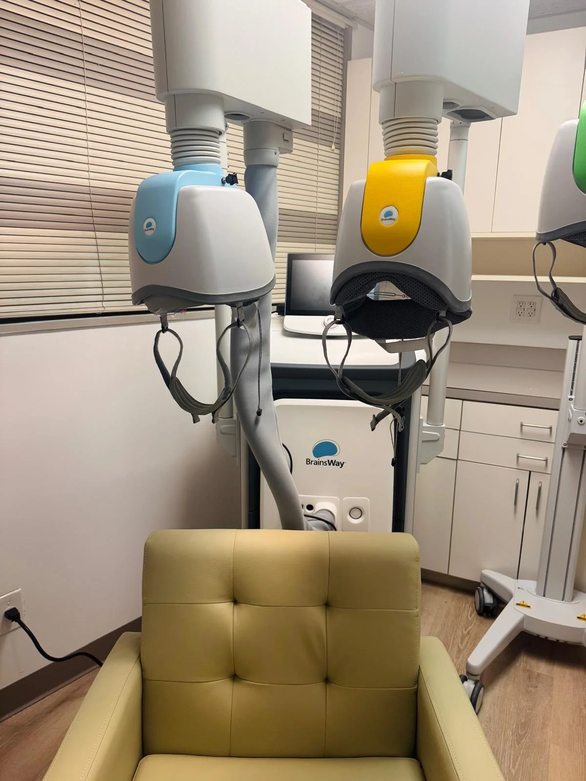 Medical ventilation equipment with two helmets color-coded blue and yellow, attached to a ventilator machine labeled 'BrainsWay', in a clinical setting with a beige sofa in the foreground.