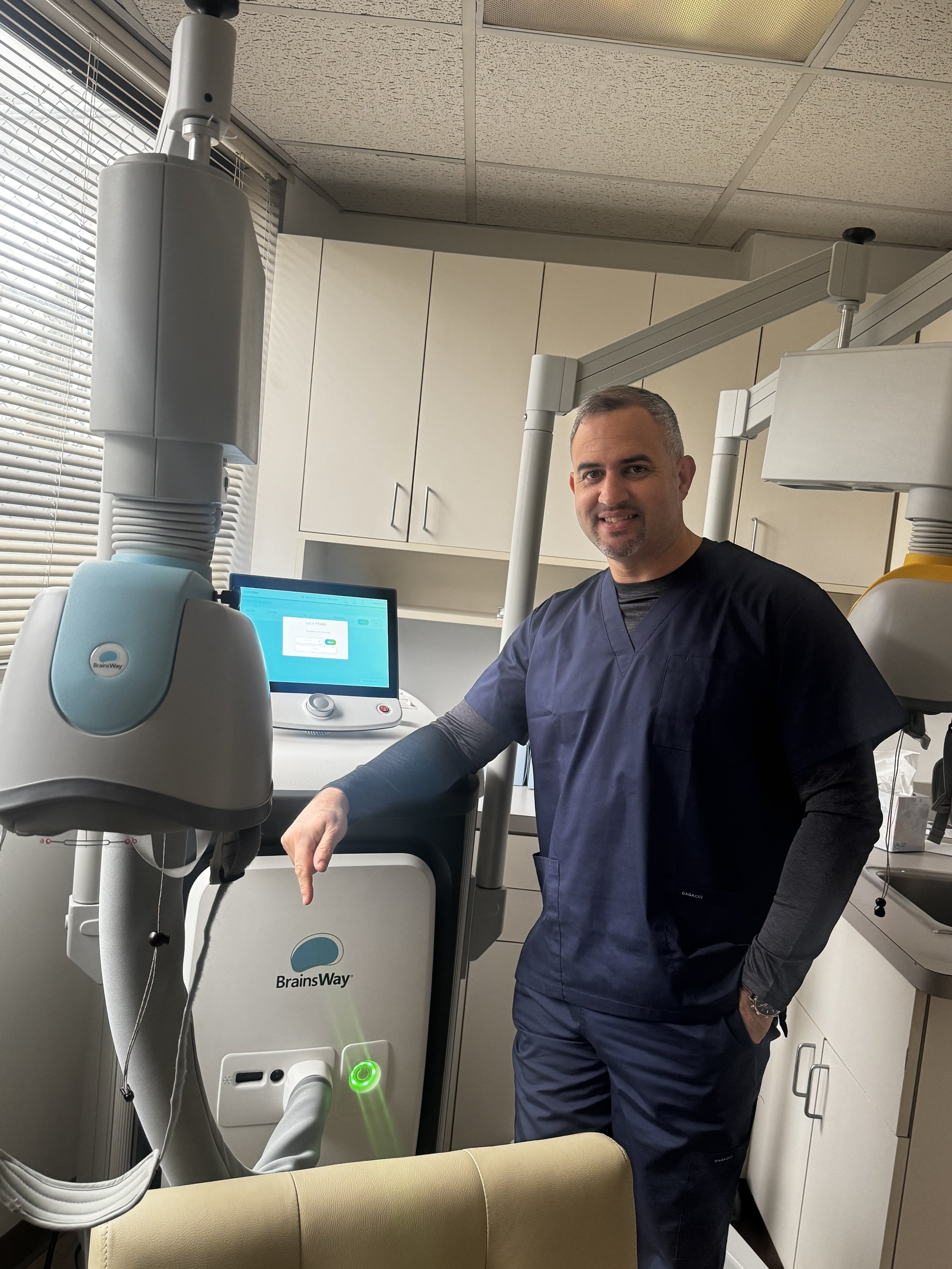 A male healthcare professional in navy scrubs standing in a medical room next to brain scanning equipment, with a computer monitor on the counter behind him.