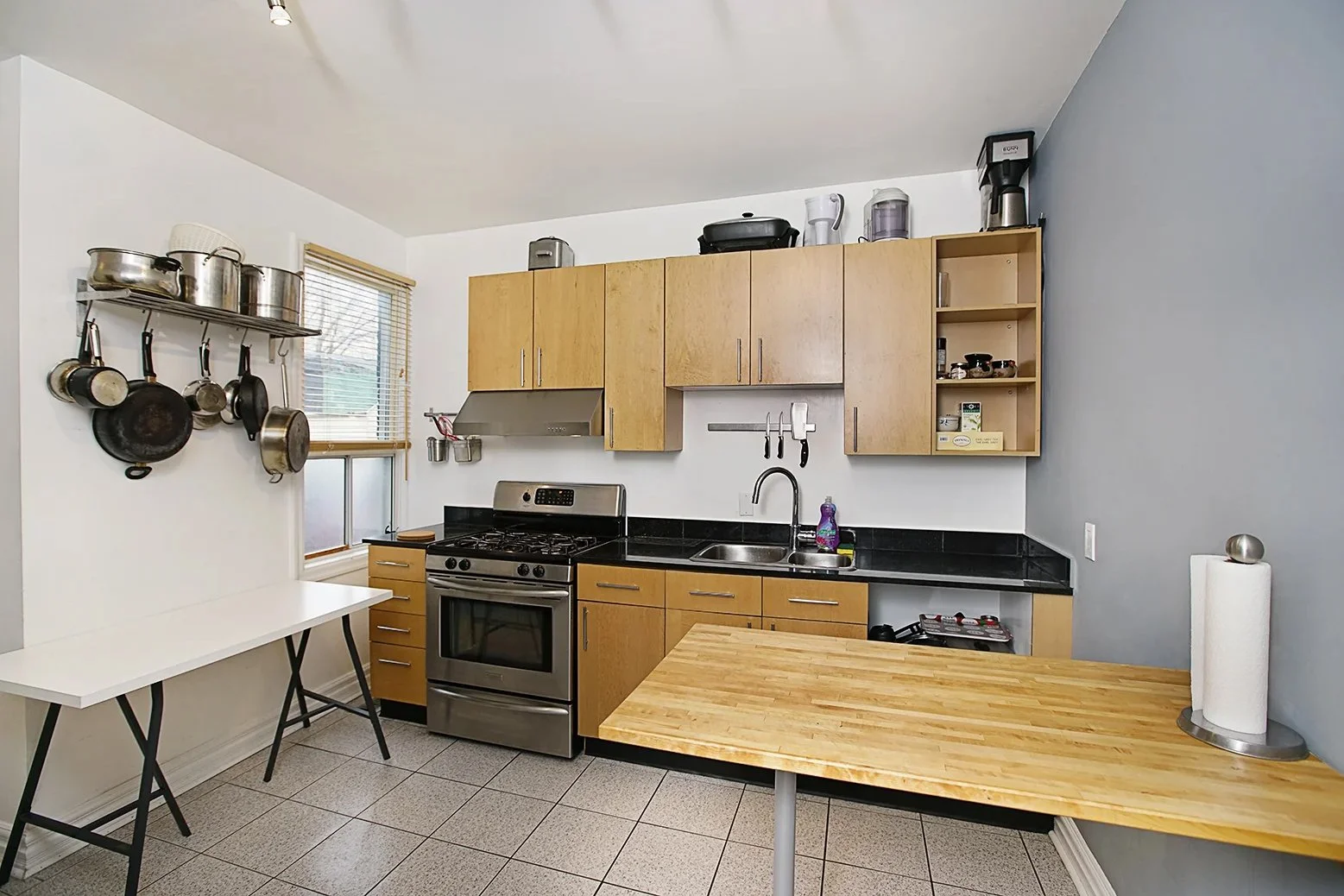 A kitchen with wooden cabinets, a silver stove, black countertop, and a stainless steel sink. Hanging pots and pans on the white wall, with a small window with blinds. A wooden table with a paper towel roll on it in the foreground.