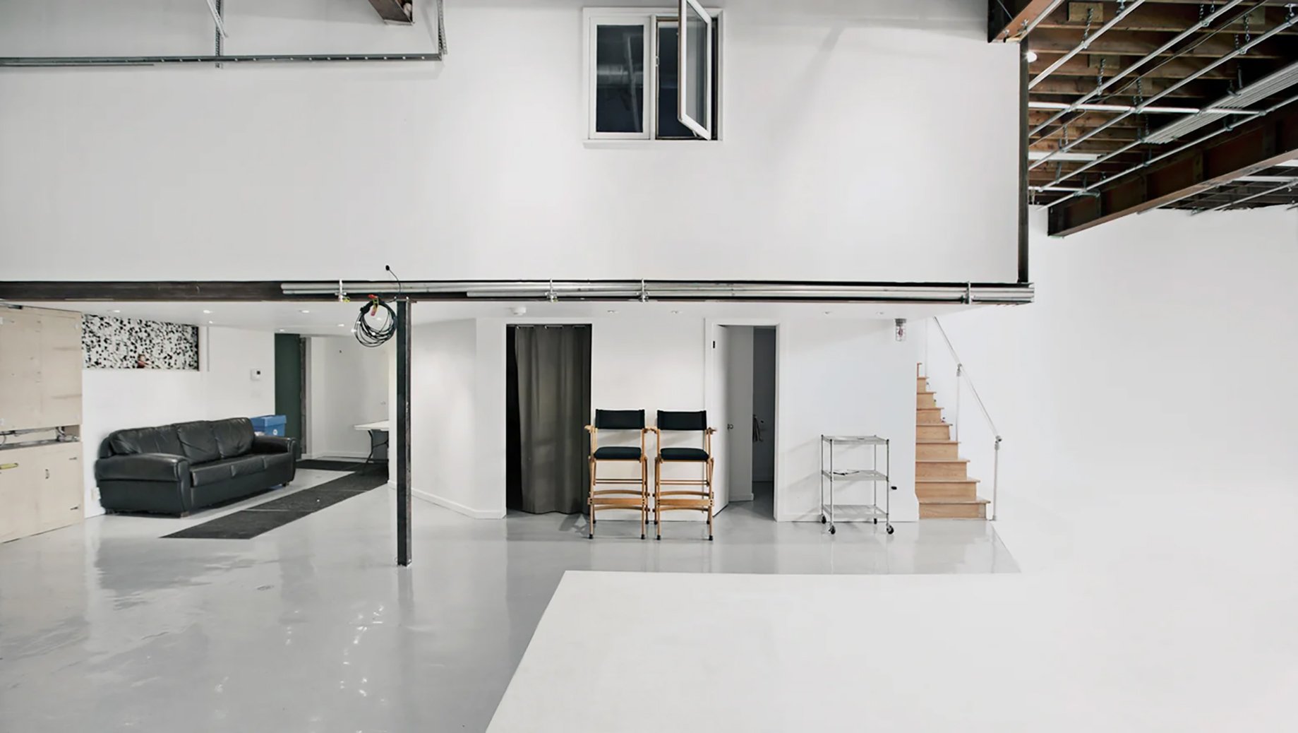 Interior of a minimalistic, industrial-style room with white walls, a black leather sofa, two black and wood chairs, a small metal cart, and a staircase with light wooden steps, in a space under construction or renovation.