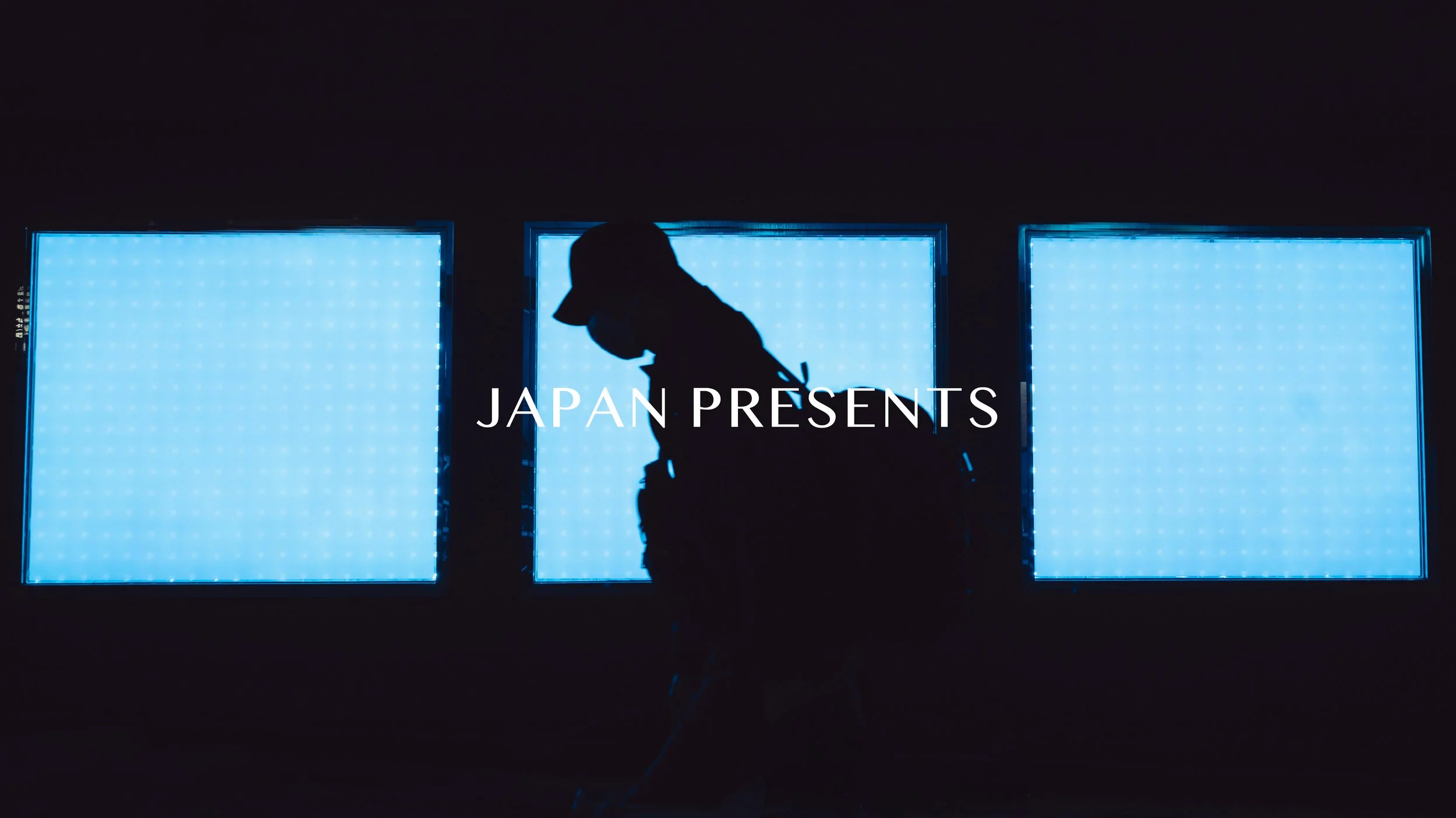 Silhouette of a person wearing a cap and glasses, standing in front of three illuminated blue display panels with the words 'JAPAN PRESENTS' overlaid.