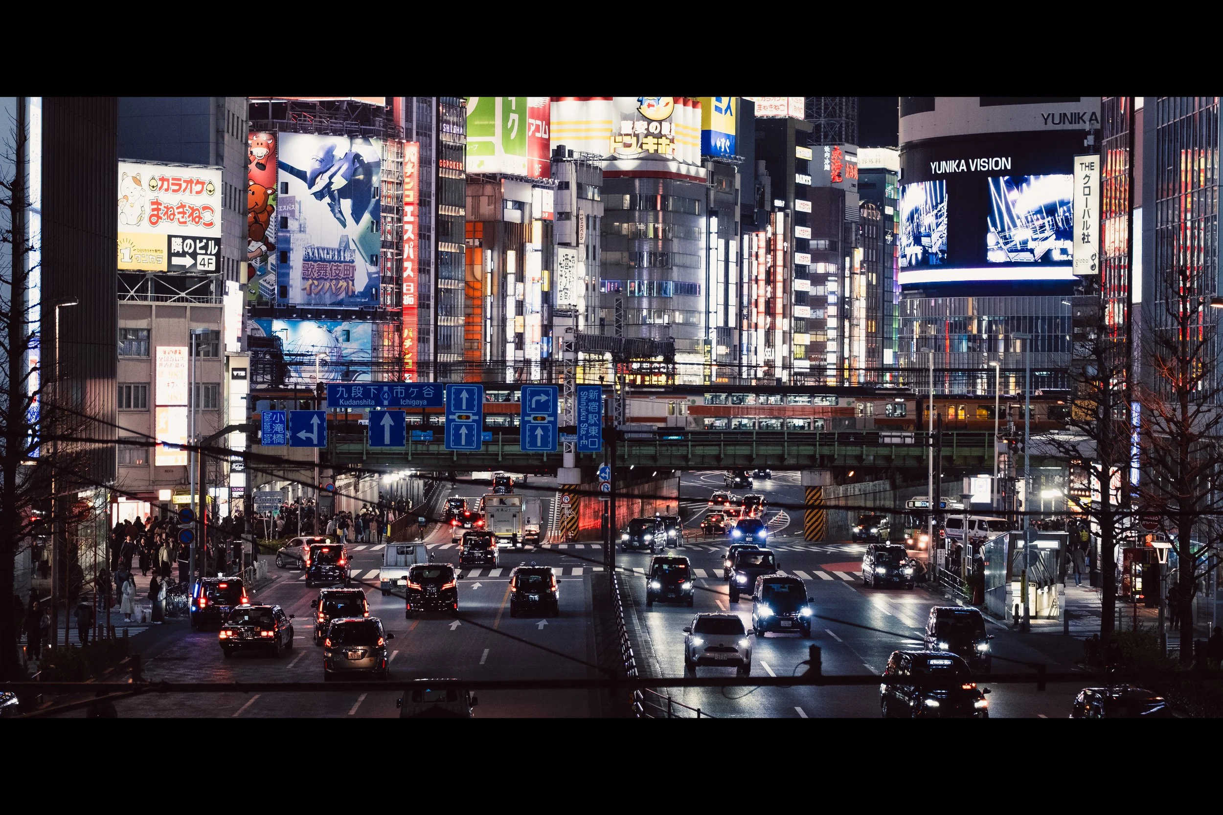 Nighttime cityscape with busy traffic, illuminated billboards, and skyscrapers with colorful lights in an urban area.