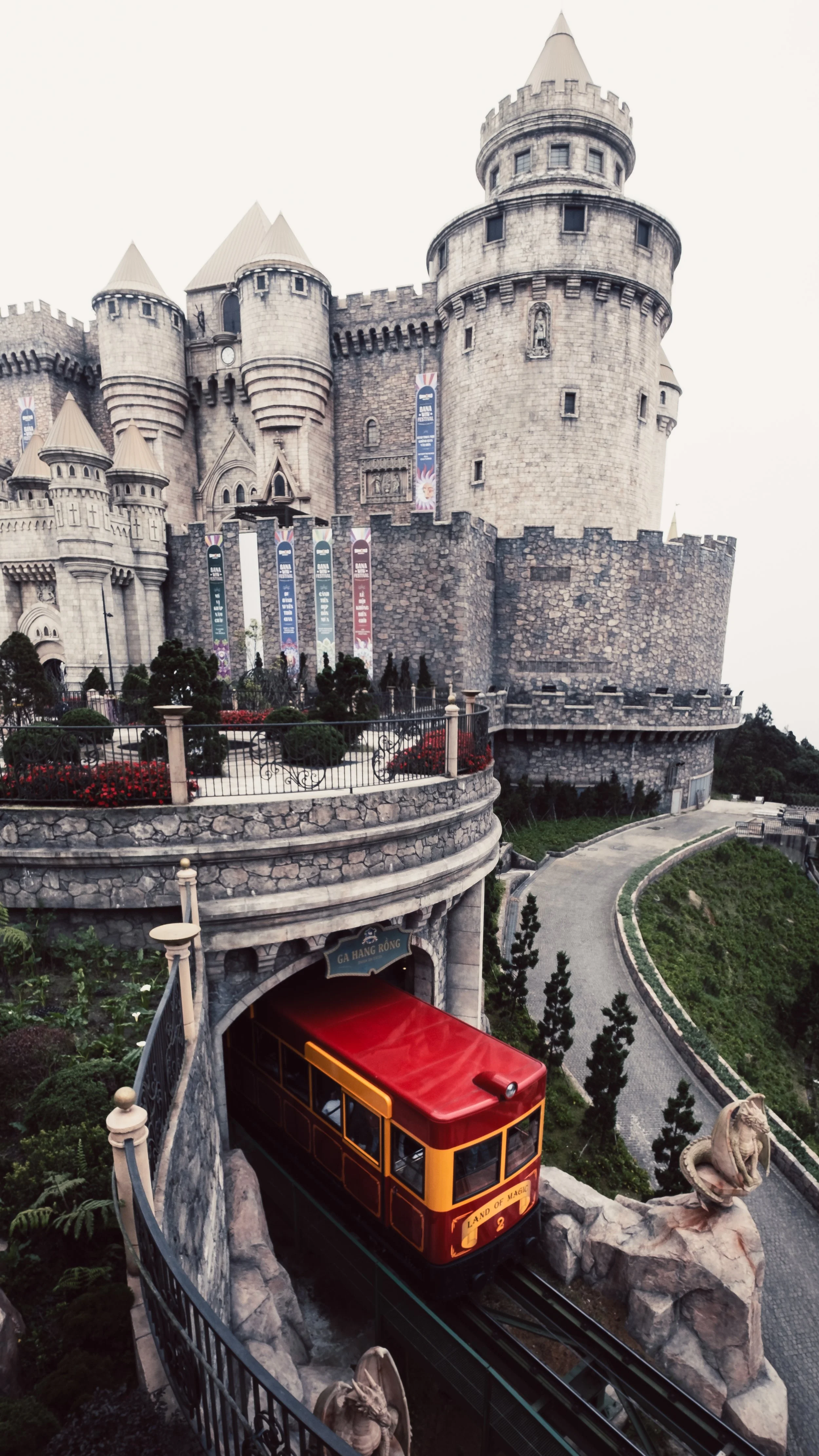 A red and yellow tram emerging from a tunnel beneath a stone castle with towers and battlements, surrounded by landscaped gardens and a curved pathway.