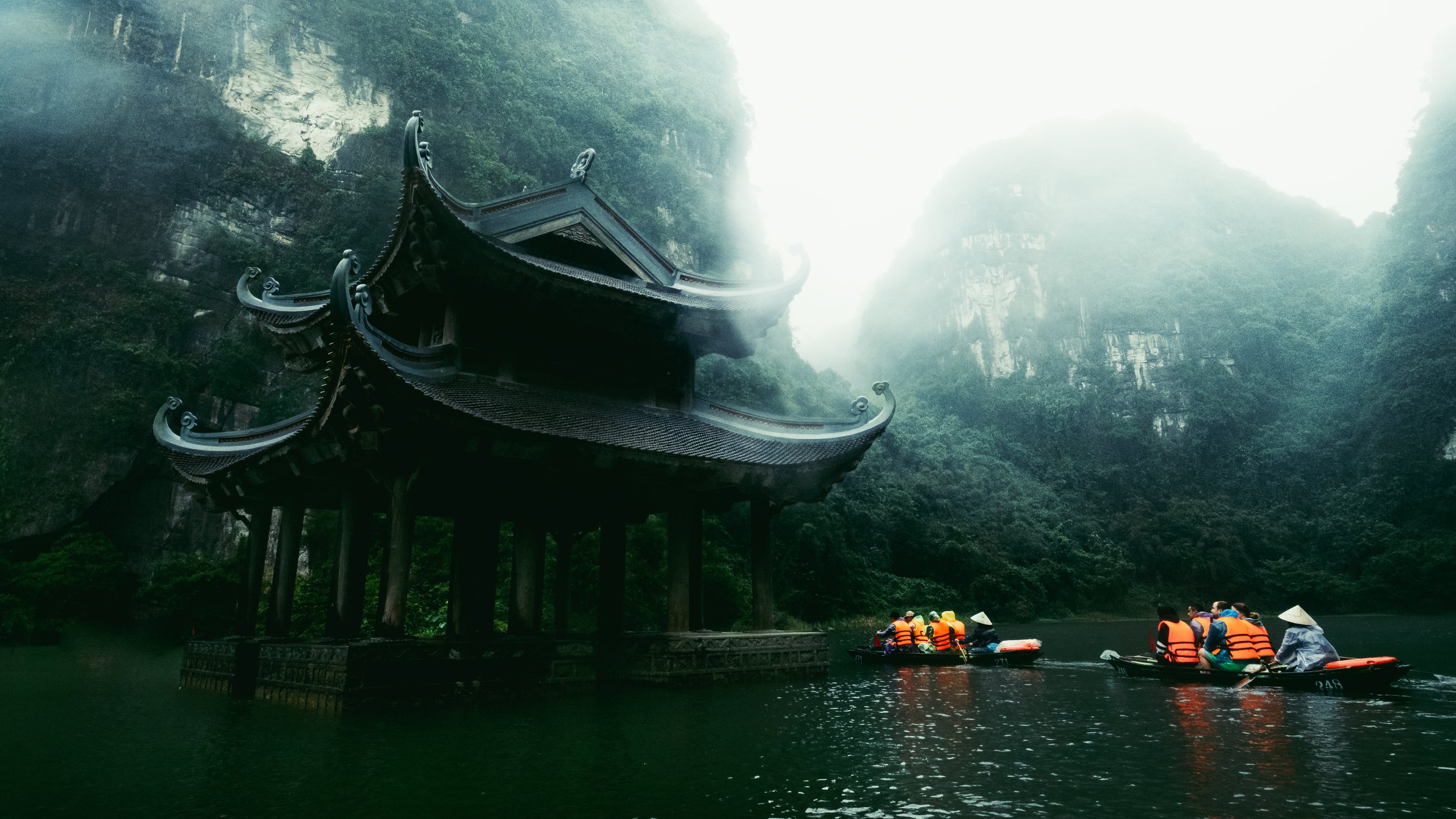 Traditional Asian pagoda located in Ninh Binh within Vietnam on stilts above water, surrounded by lush green mountains, with boaters wearing orange life jackets in the river.