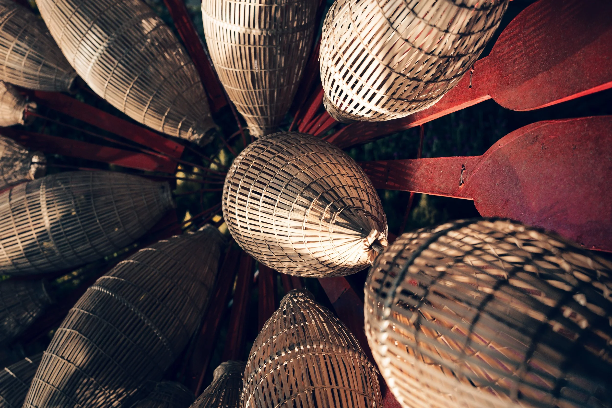 A close-up from below of traditional bamboo fish traps or conical straw containers hanging from a red metal structure, with sunlight illuminating the woven bamboo patterns.