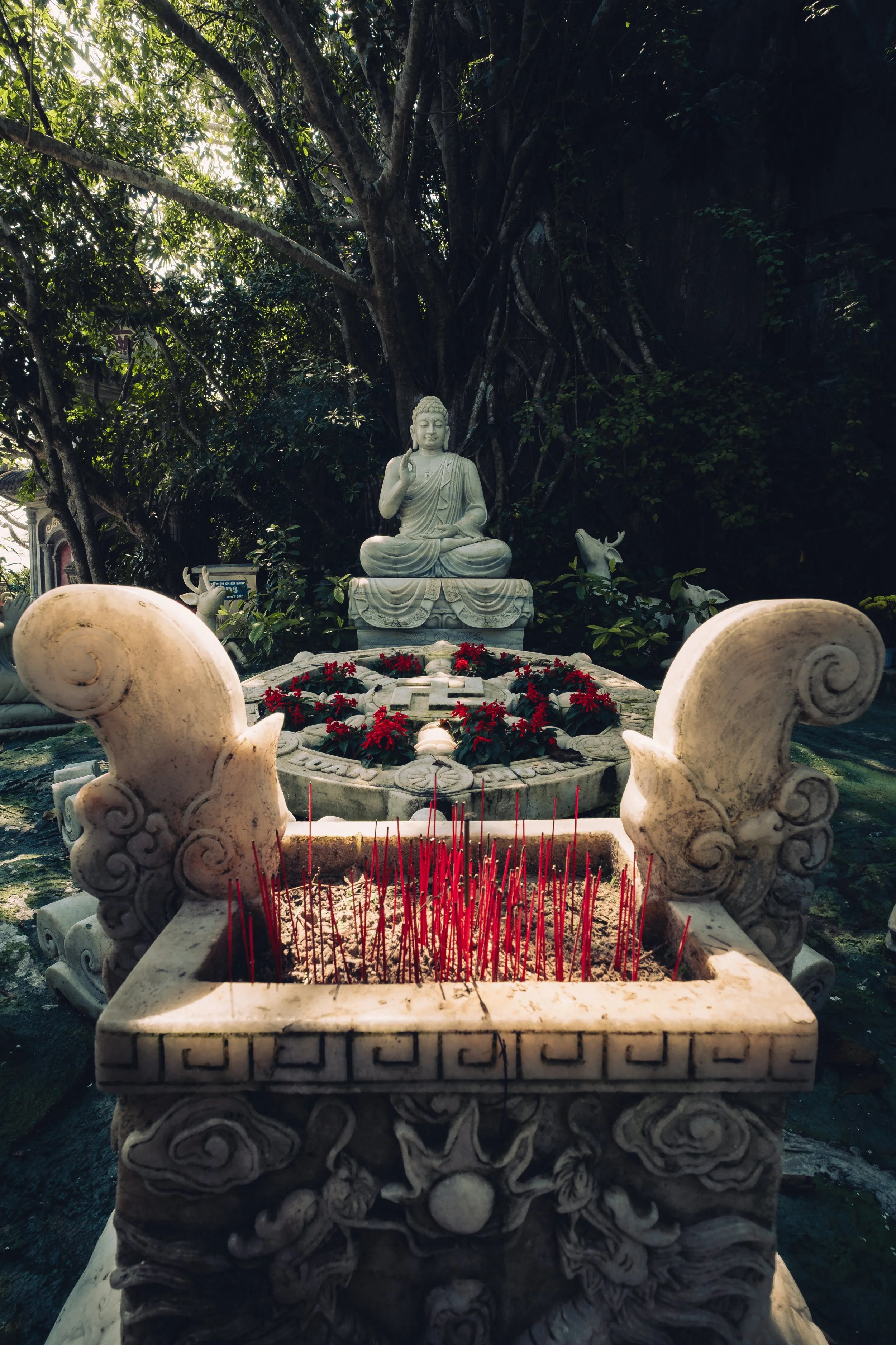 A Buddha statue outdoors behind a fountain with red flowers. In the foreground is a stone incense holder with red incense sticks.