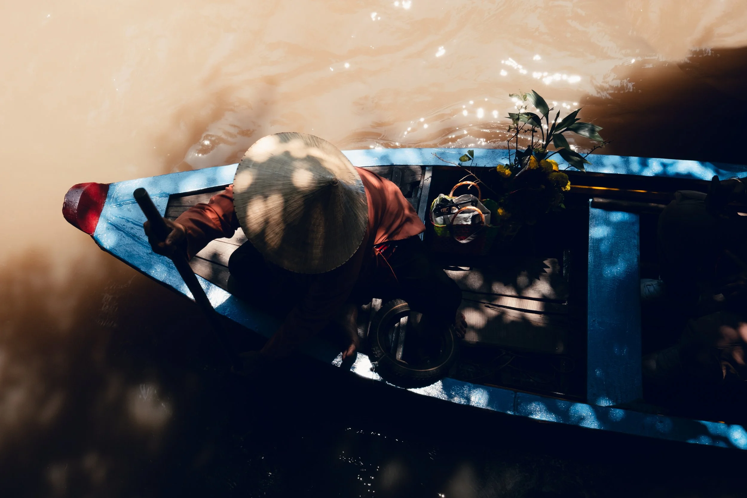 Top-down view of a person wearing a conical hat on a small boat with a bouquet of flowers and a basket, floating on muddy water.