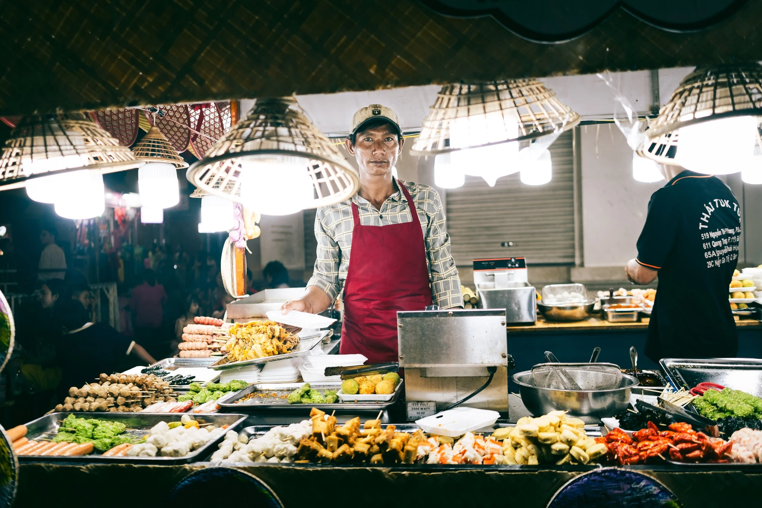 A street food vendor wearing a red apron and a cap stands behind a stall filled with various grilled and fried foods at night, illuminated by hanging lamps.