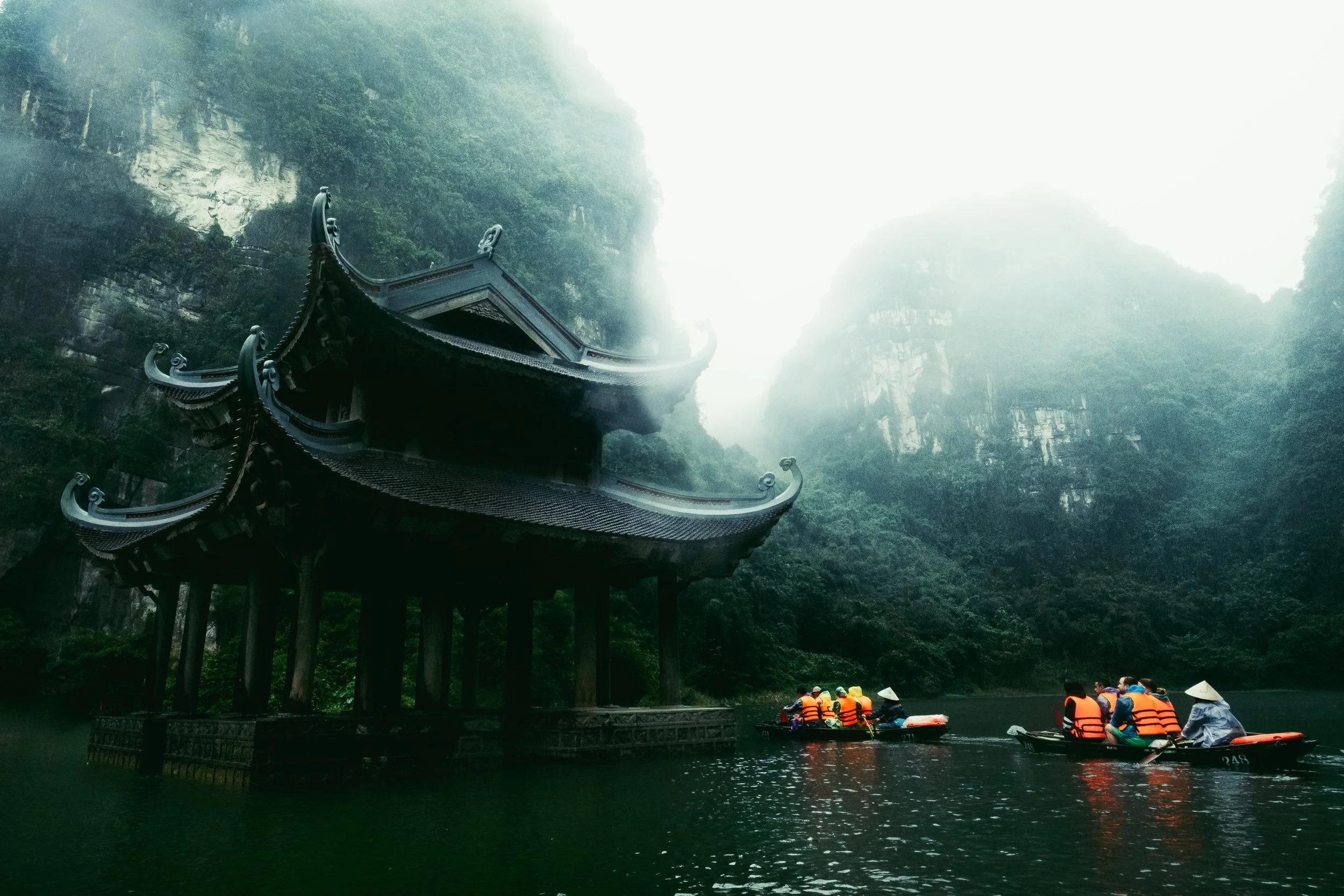 Traditional Chinese pavilion on water with lush green mountains and mist in the background, several people in boats wearing life jackets and conical hats.
