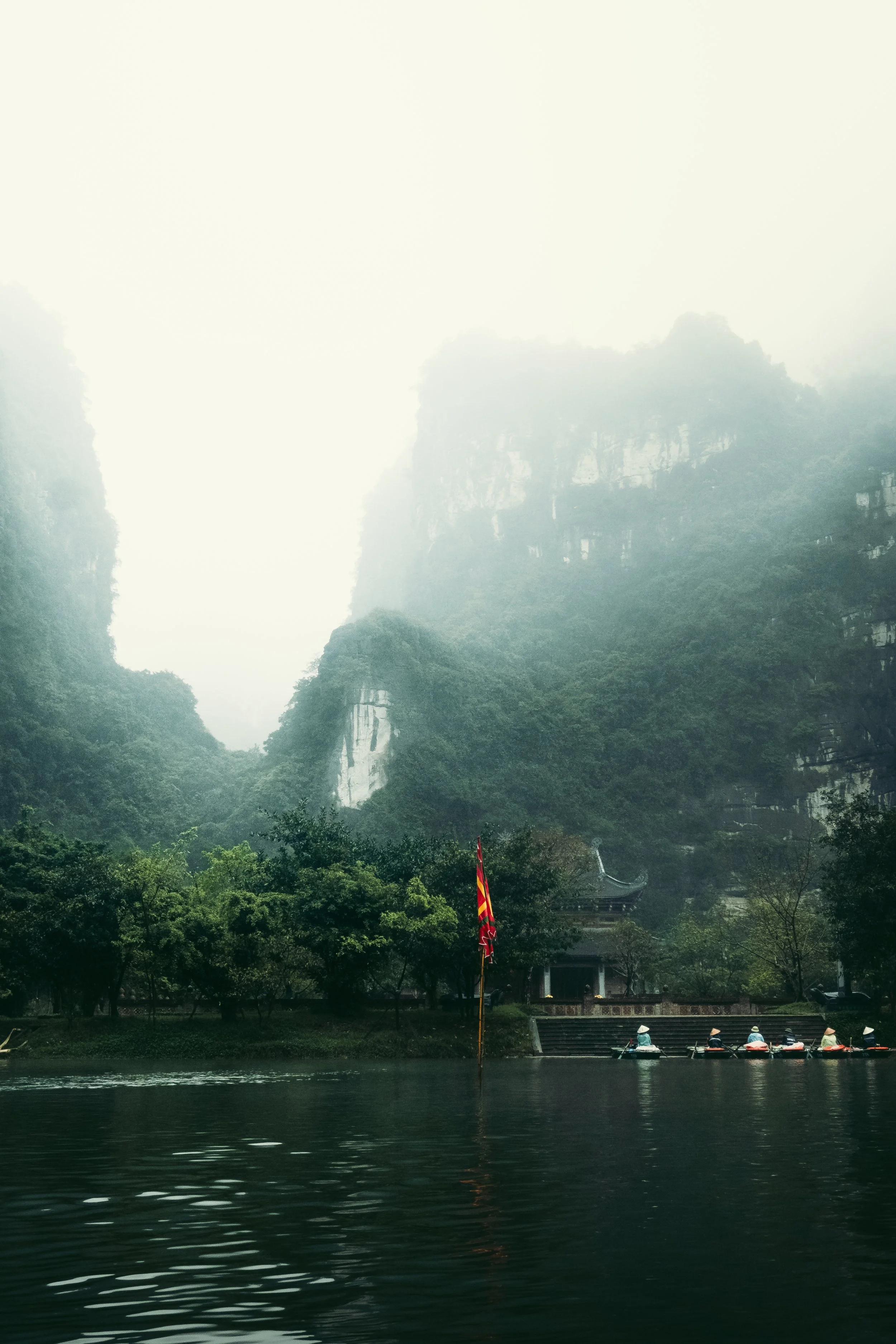 Mountains covered in mist over a calm river with a small boathouse, trees, and a flag on a pole.