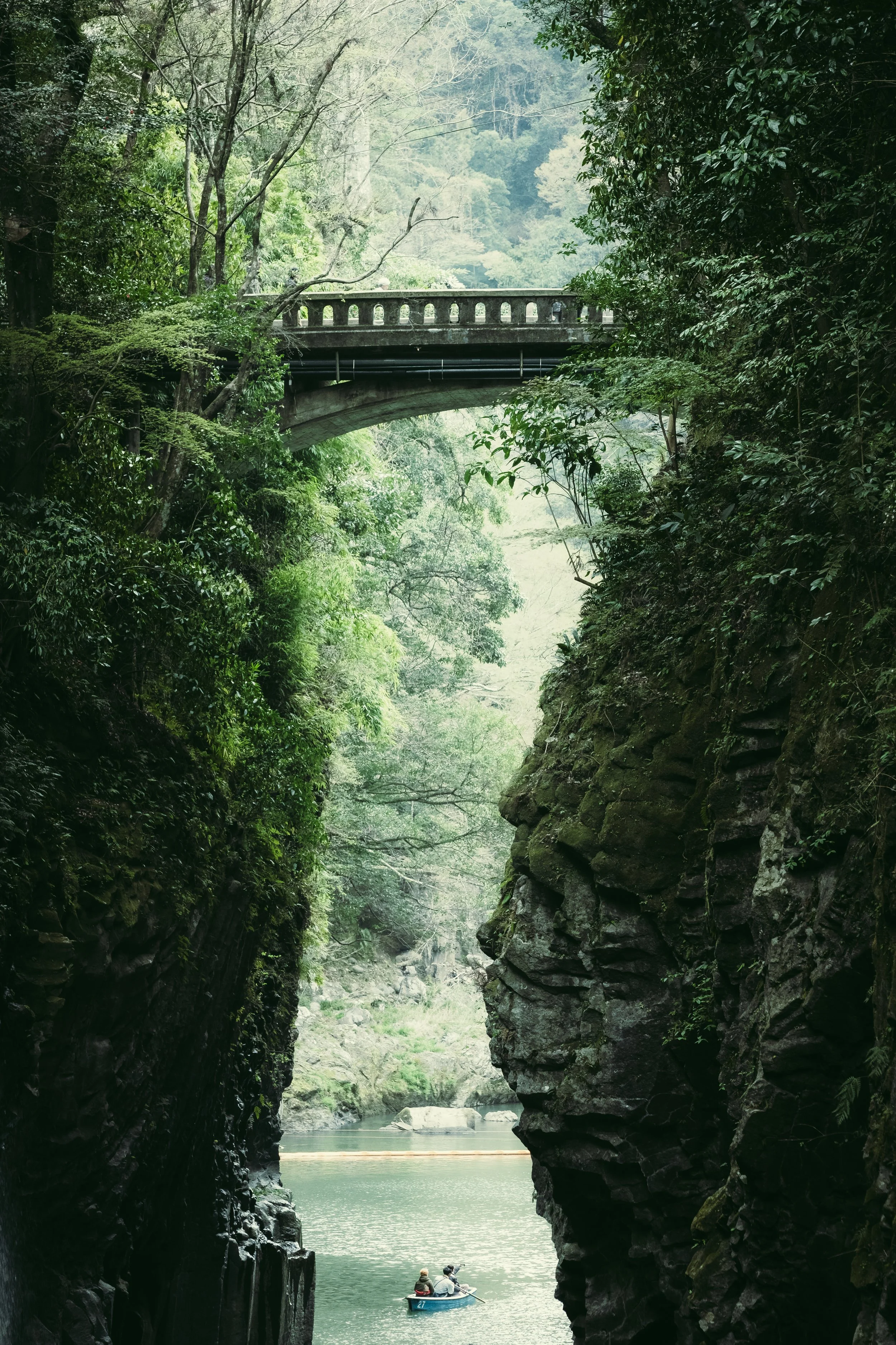 A person canoeing in a narrow, rocky gorge with lush green trees and a bridge overhead.