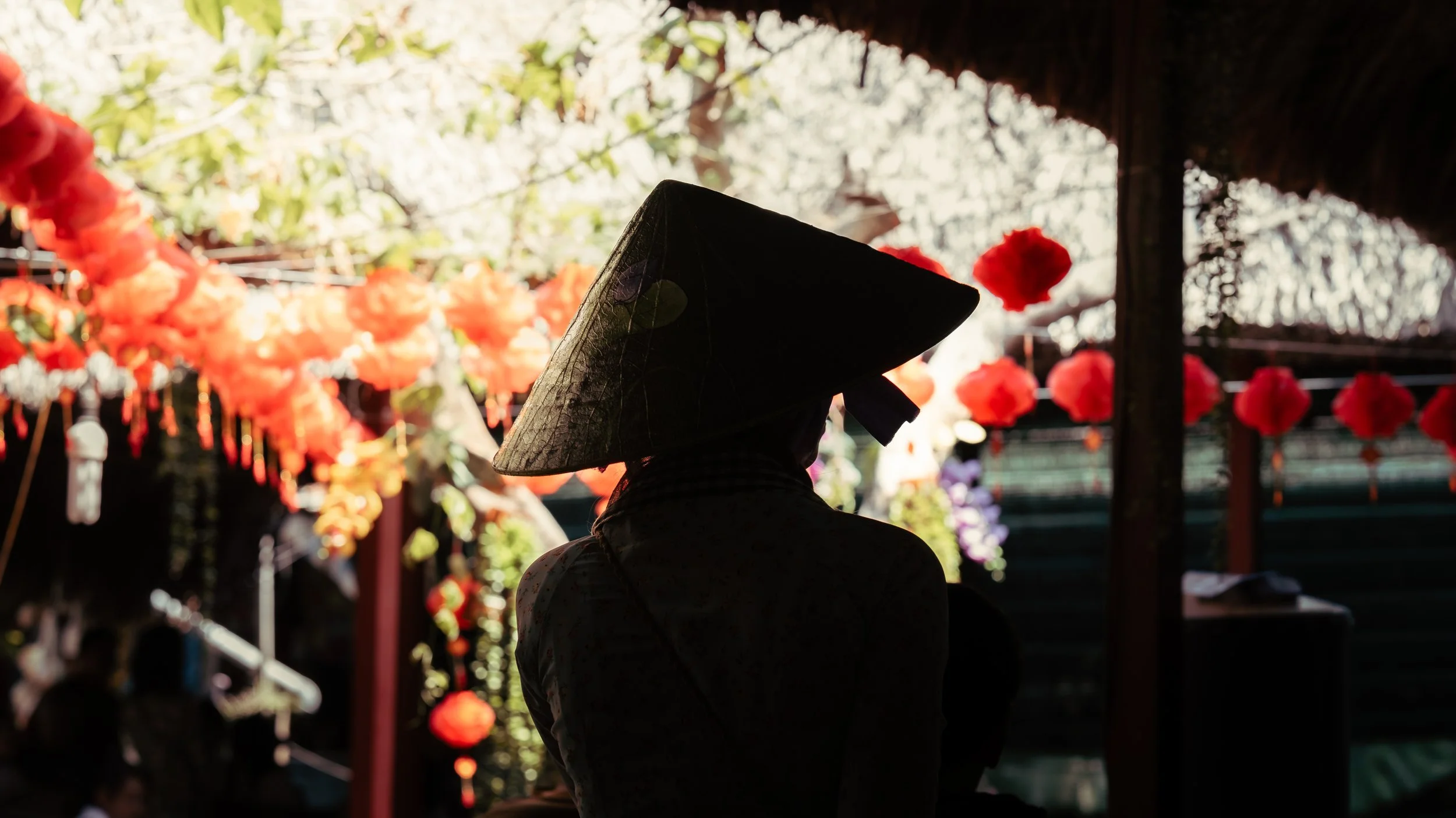 Silhouette of a woman wearing a traditional Asian conical hat, standing under red lanterns and decorations, backlit by bright outdoor light.