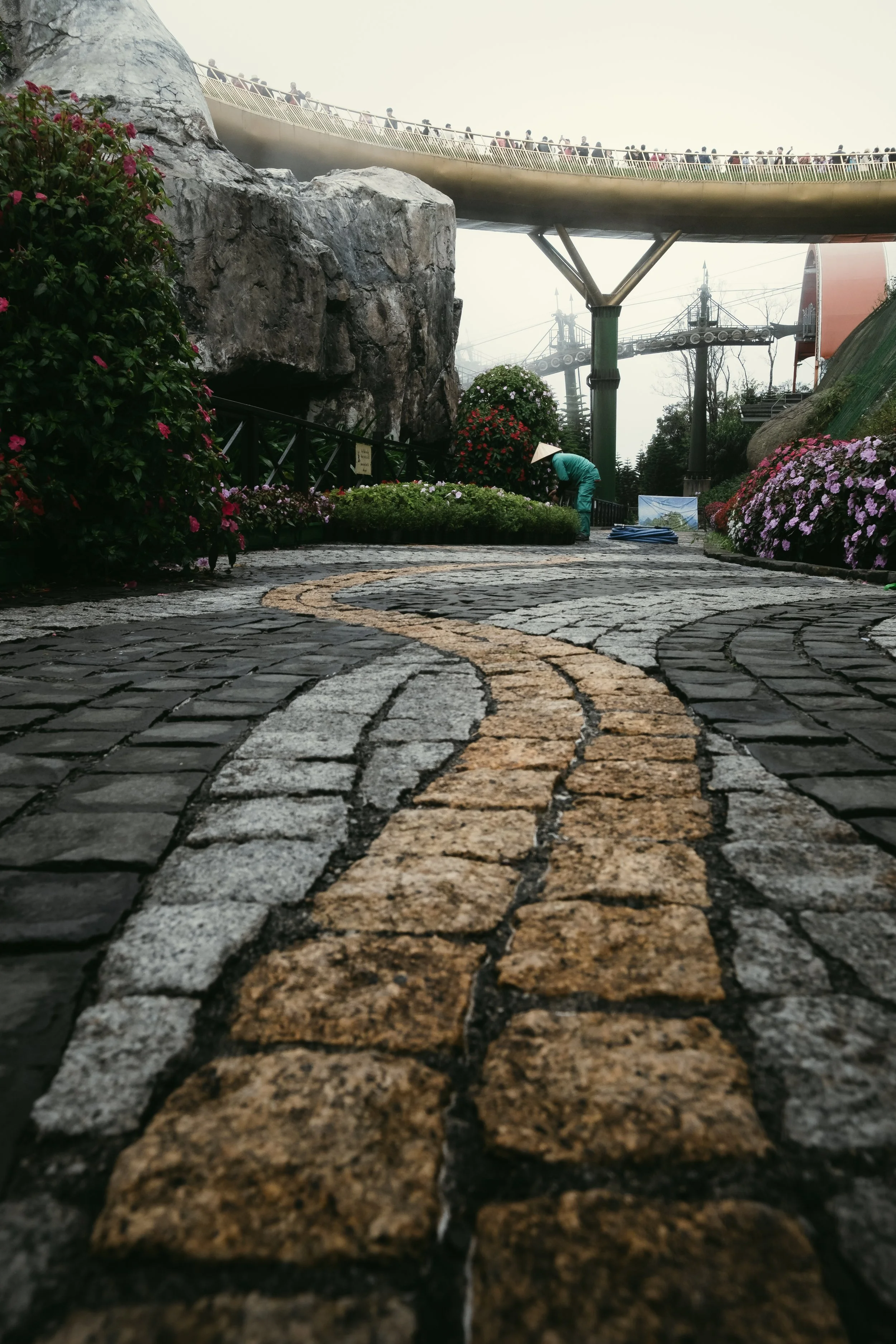 A cobblestone pathway with a yellow line in the center, outdoor park with colorful flowers, large rock formations, a person in a turquoise outfit and conical hat tending to plants, and a curved bridge or walkway overhead.
