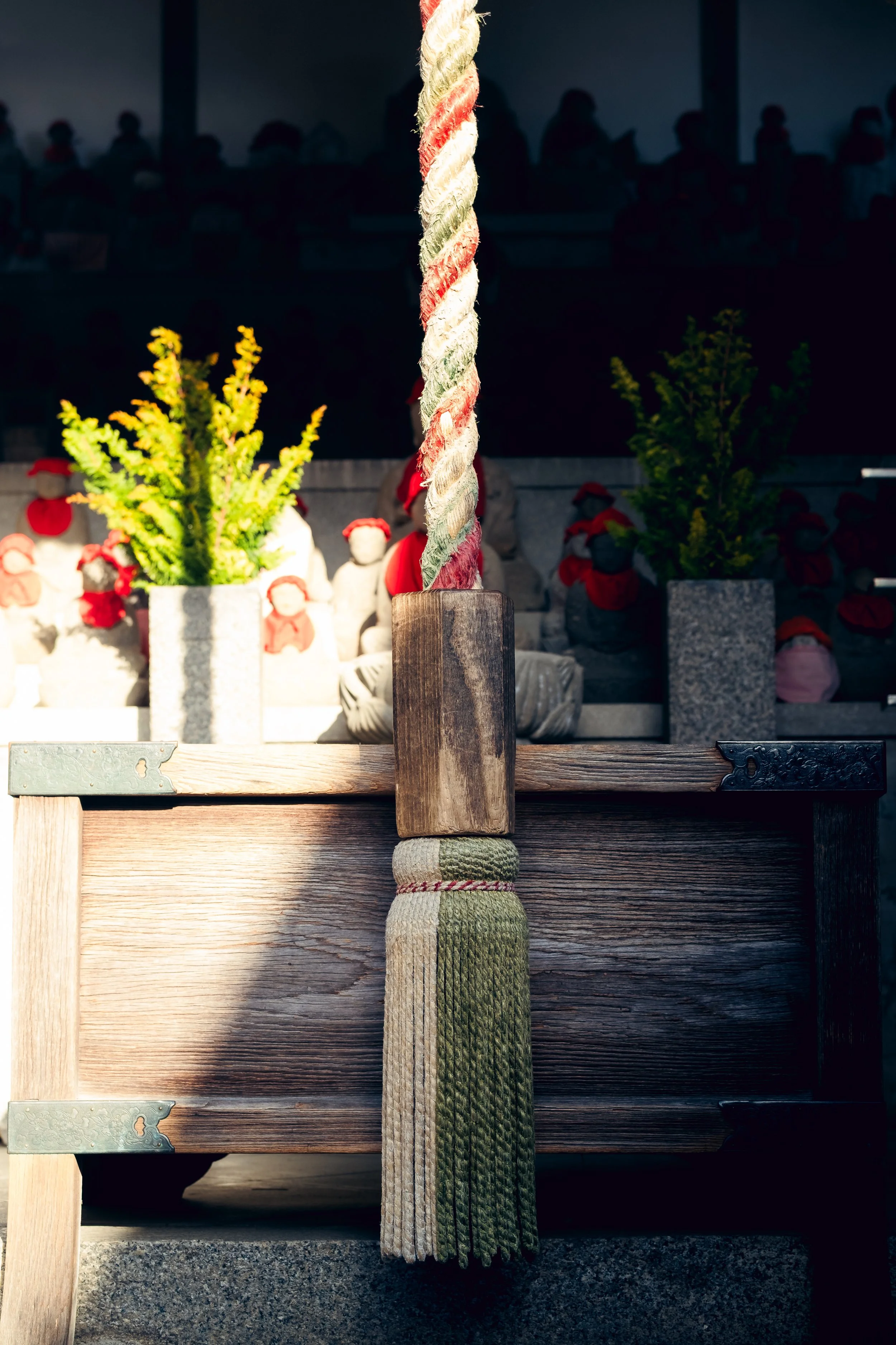 Close-up of a decorative shimenawa rope with paper streamers, hanging over a wooden structure at a Japanese shrine, with stone statues and potted plants in the background.