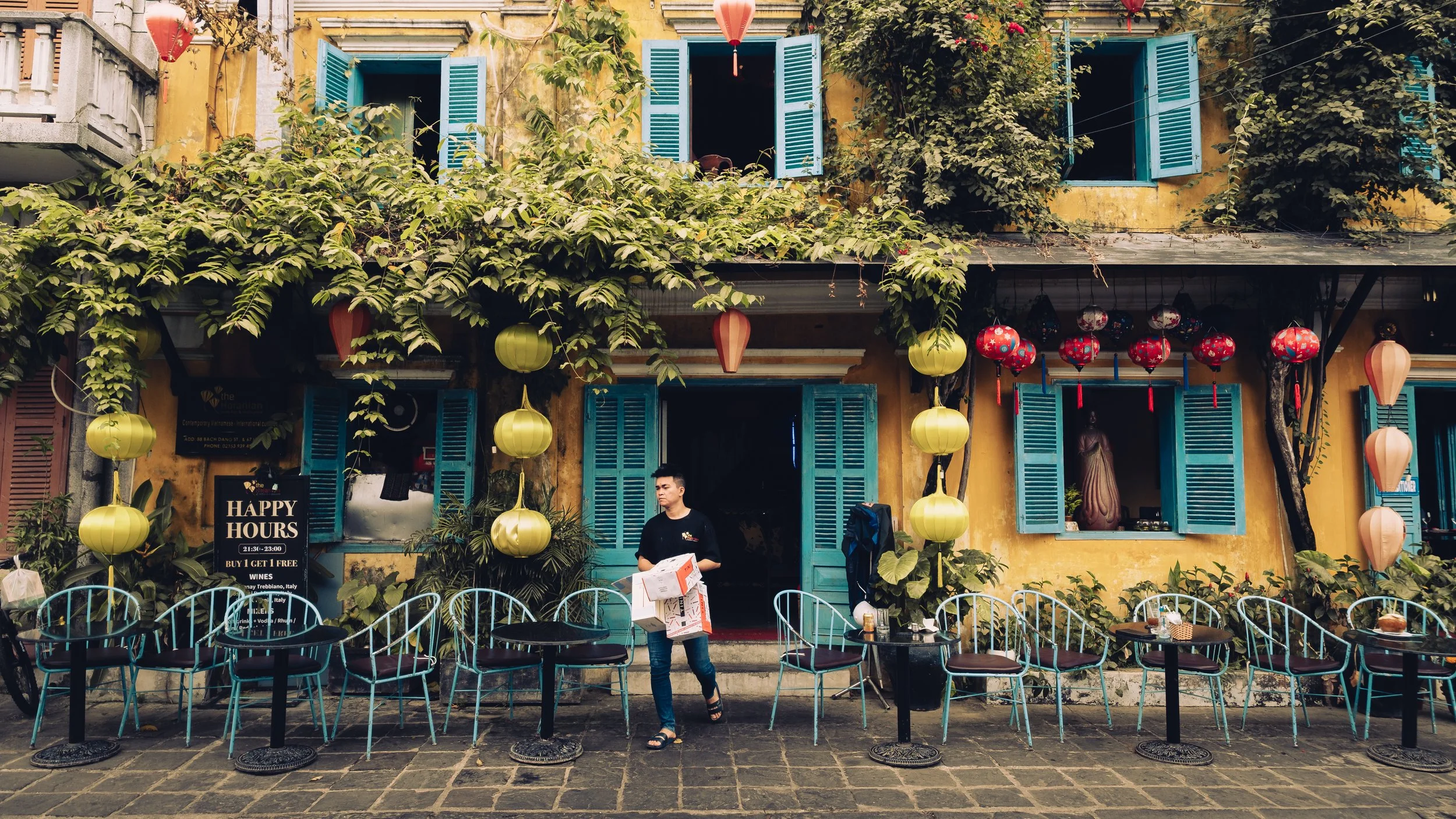 Yellow building with open blue shutters and red and green hanging lanterns, outdoor seating with black tables and blue chairs, and a person walking by holding newspapers.
